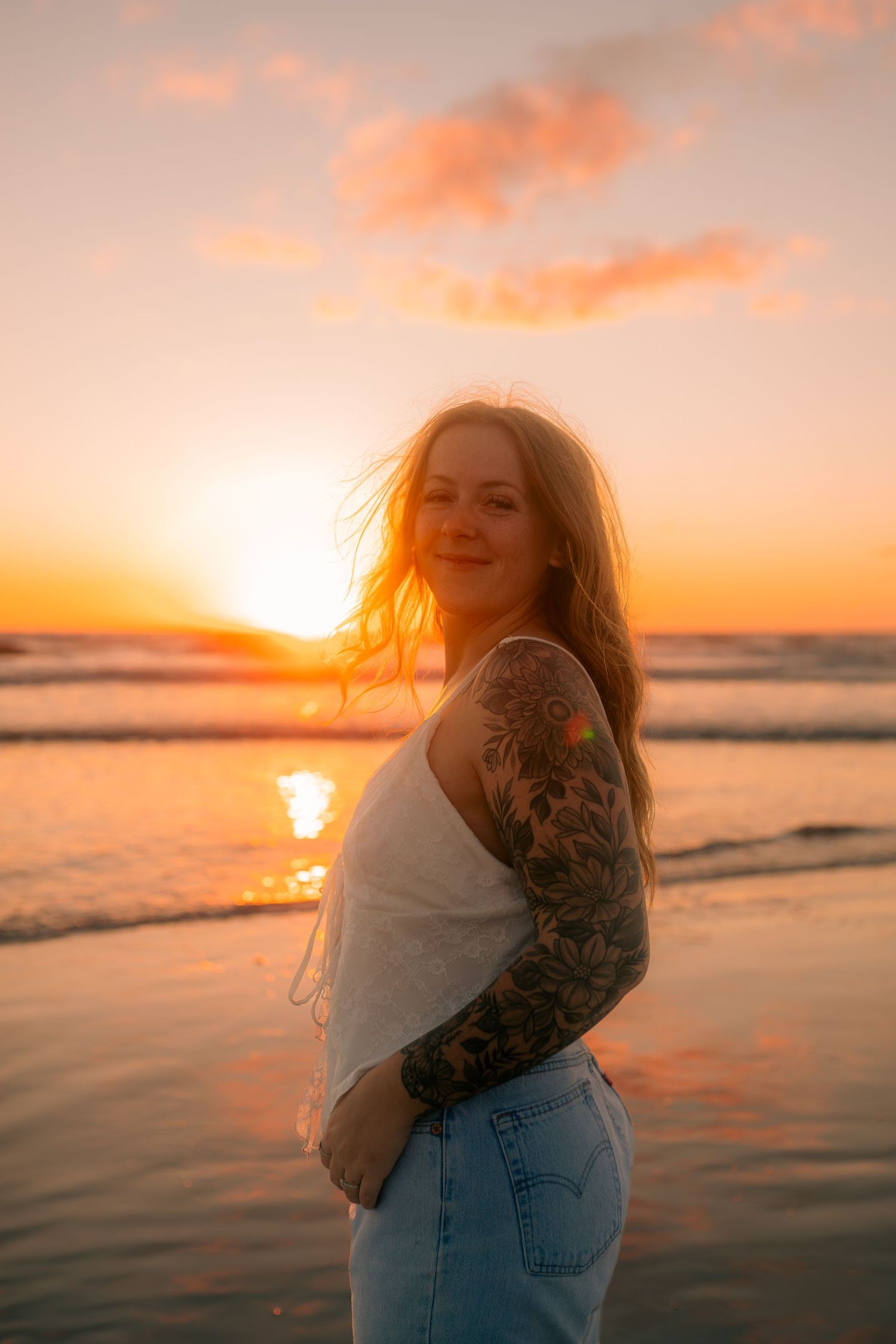 Smiling woman with tattoo sleeve on beach at golden sunset, wearing white top and jeans, ocean waves in background.