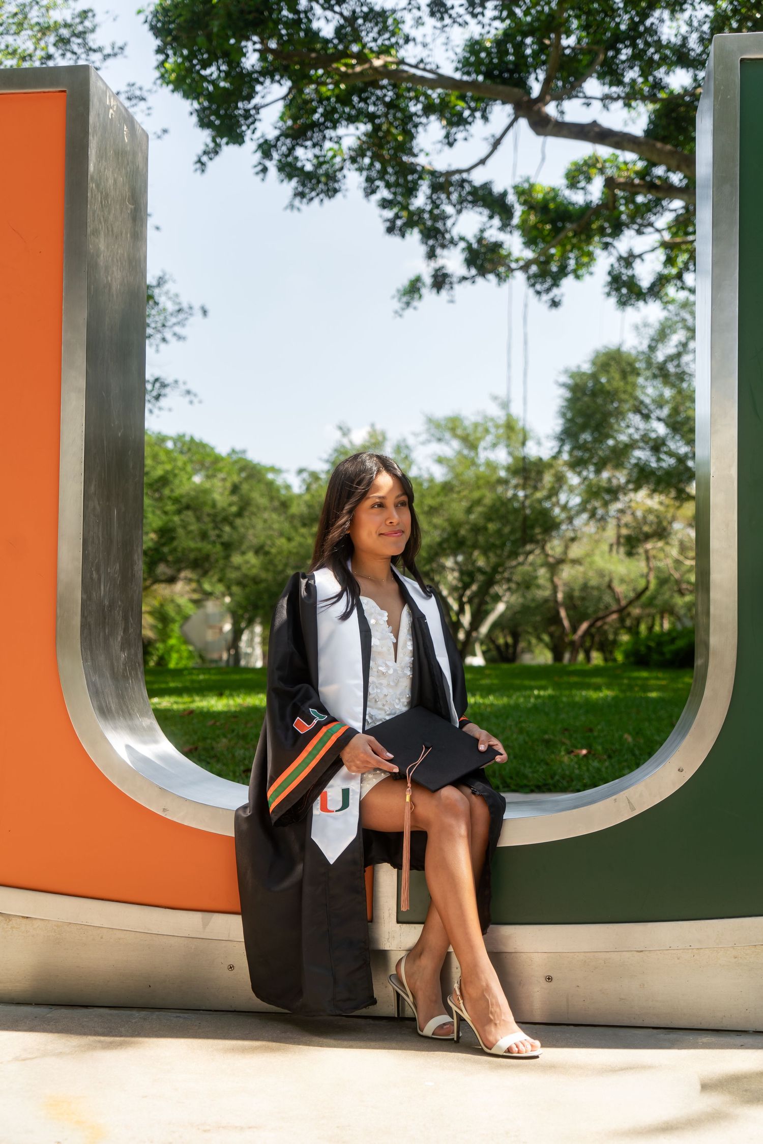 Graduate in University of Miami regalia sits holding cap beside large U-shaped campus sculpture.