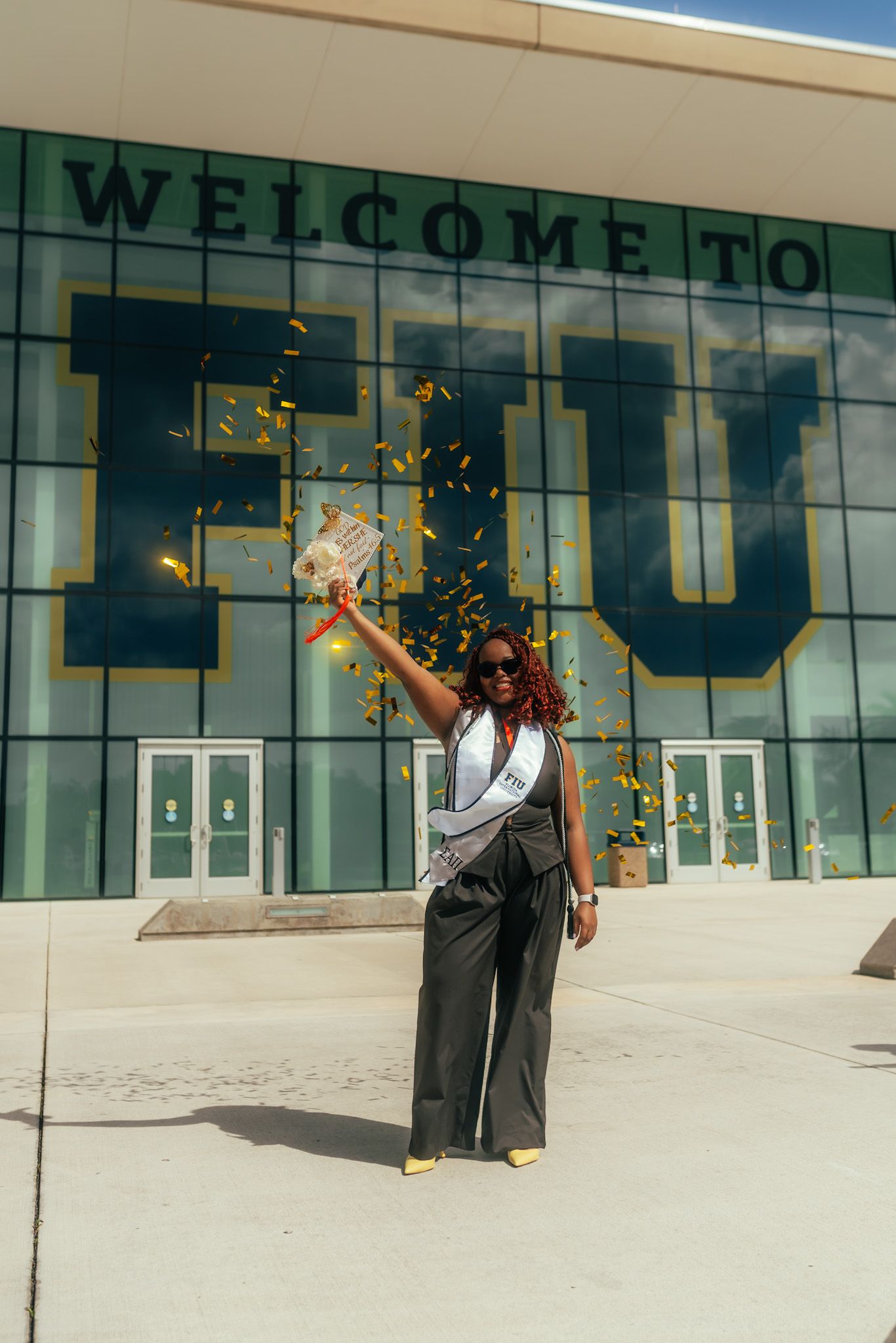 Woman celebrating with confetti in front of FIU Welcome sign, wearing sash and sunglasses outdoors.
