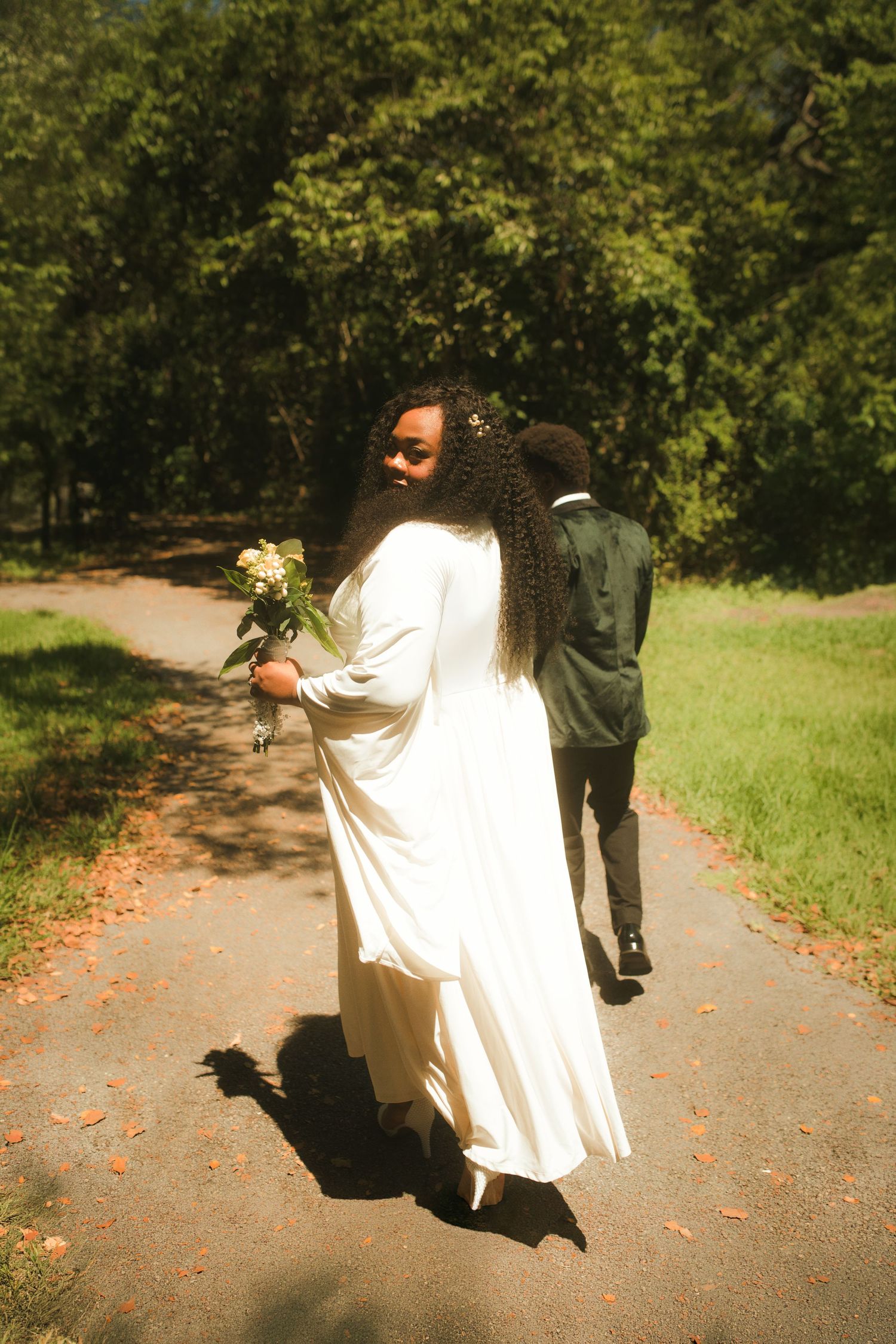 Bride in white gown holding bouquet walks with groom on tree-lined path turning to smile.