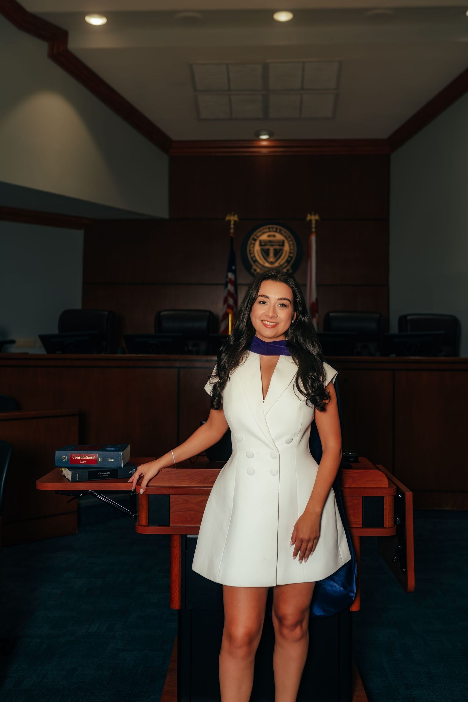 Woman in white dress with graduation stole poses in wood-paneled courtroom setting.