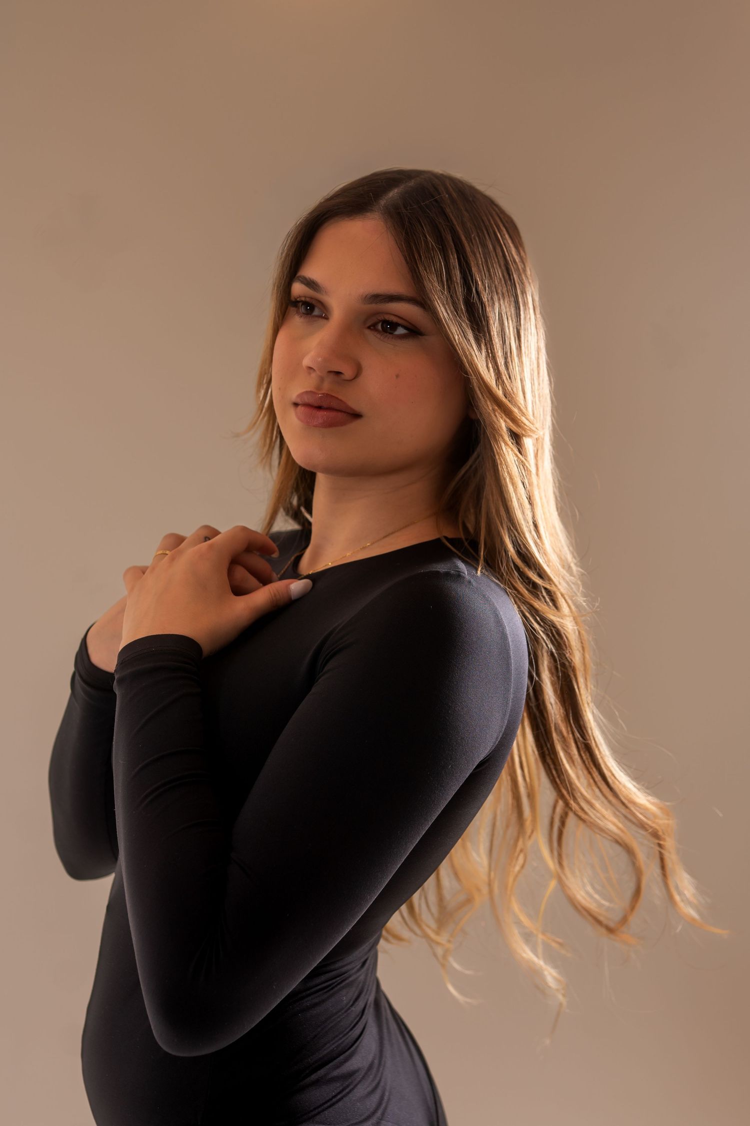 Young woman with long blonde hair poses in a black long-sleeve bodysuit against a neutral background.