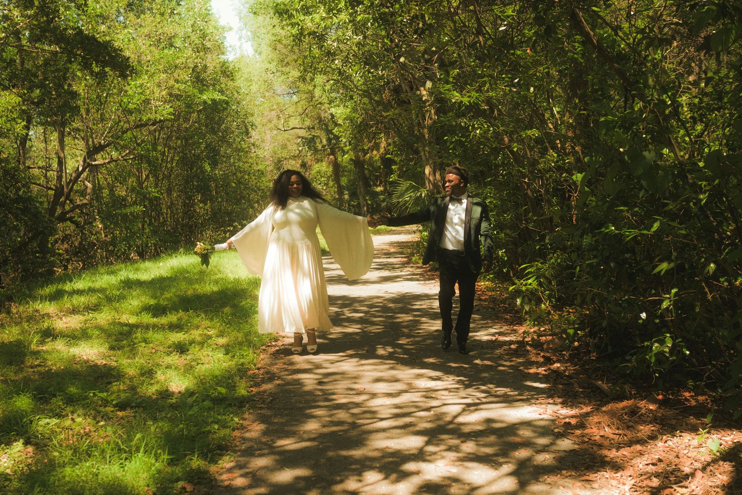 Joyful newlywed couple walking hand-in-hand on sunlit forest path in wedding attire.