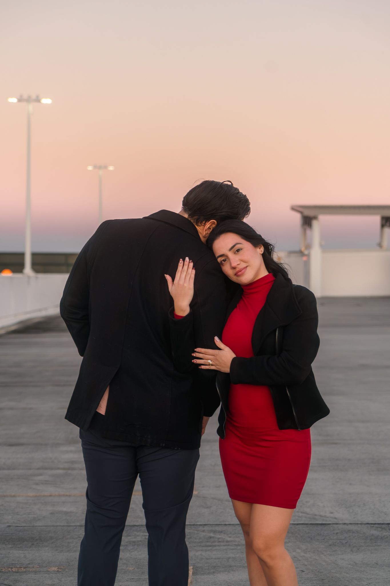Romantic couple hugging at dusk on a rooftop parking lot, woman in red dress and black jacket.