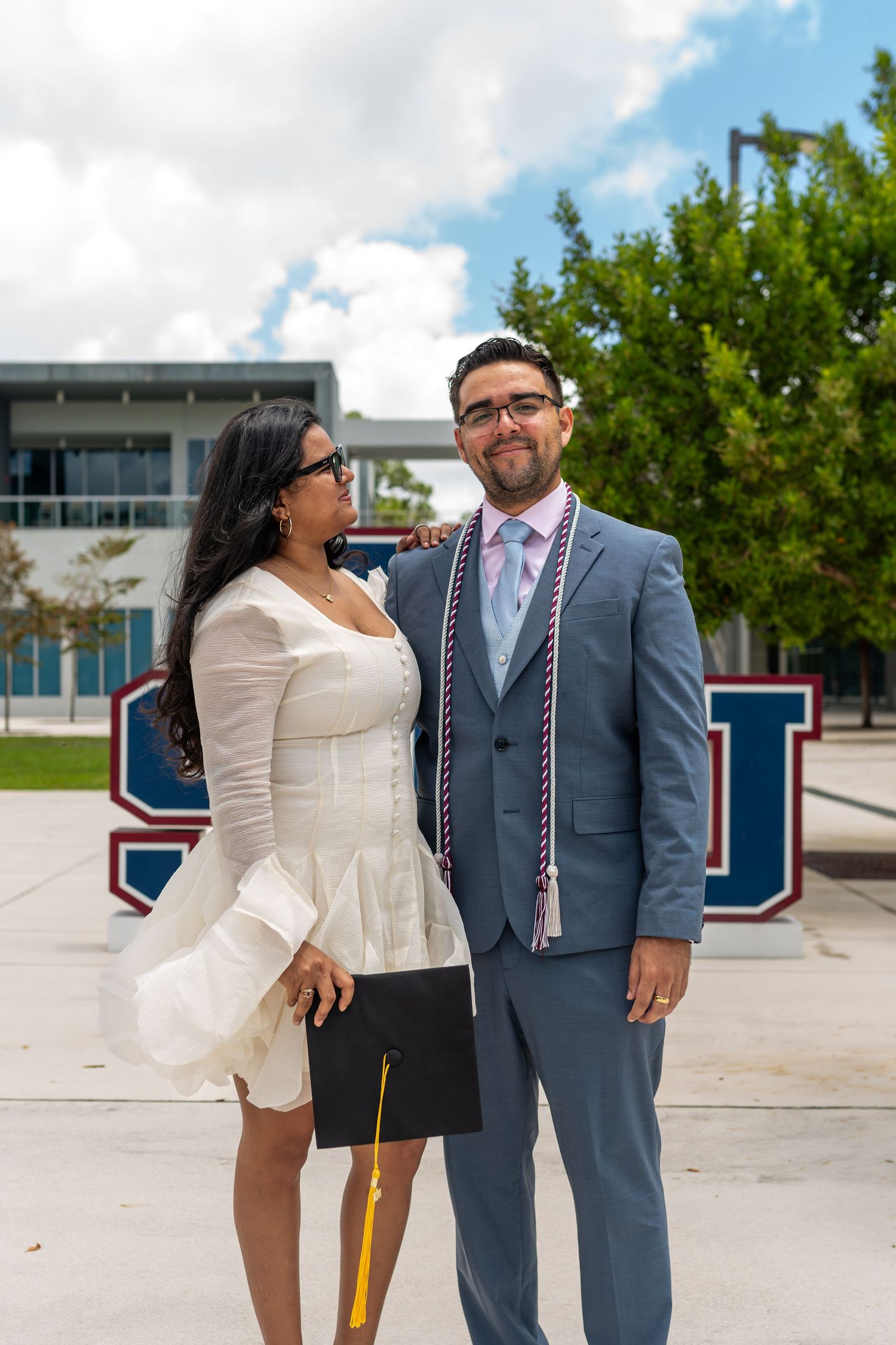 A graduate in a gray suit with stoles poses with a woman in white near an SU sign outdoors.