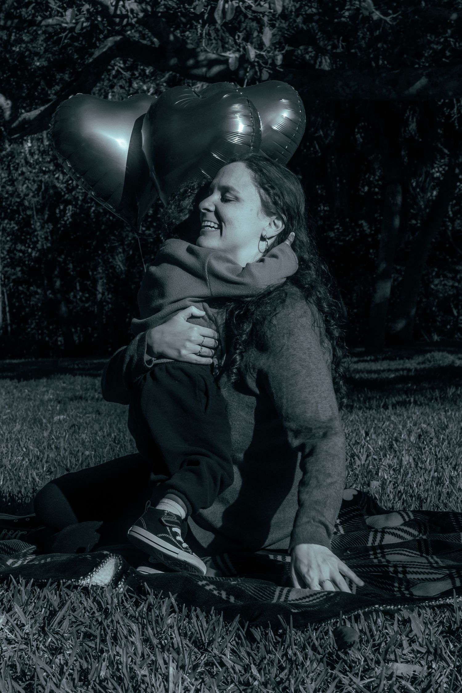 Woman smiling while hugging a child outdoors on a blanket with heart-shaped balloons in a dark toned photo.