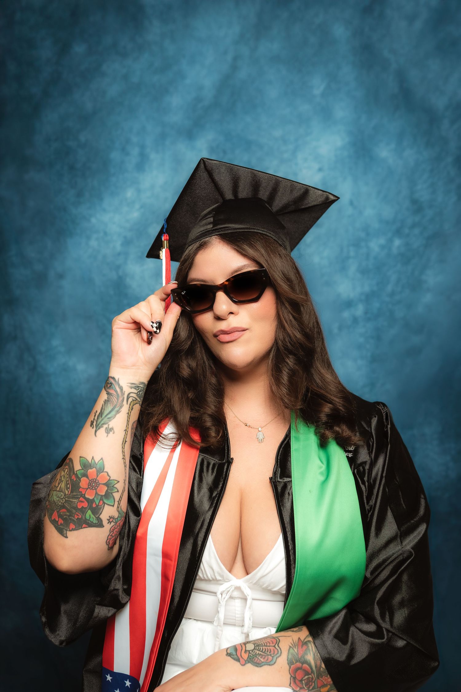 Woman in graduation cap, gown, sunglasses, and American flag sash posing confidently with tattoos visible.