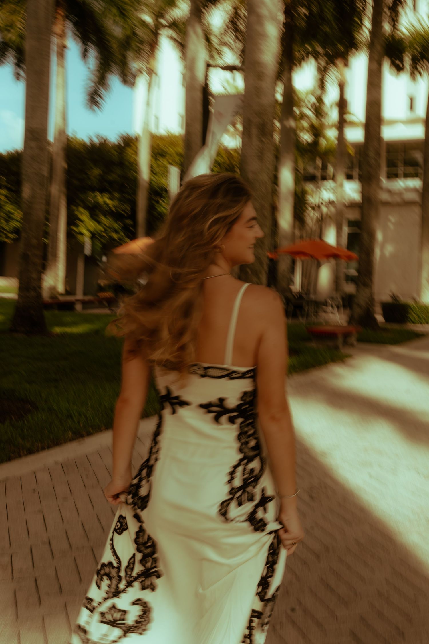 Woman in white floral maxi dress walking along tropical palm-lined path, hair flowing, captured in motion blur.