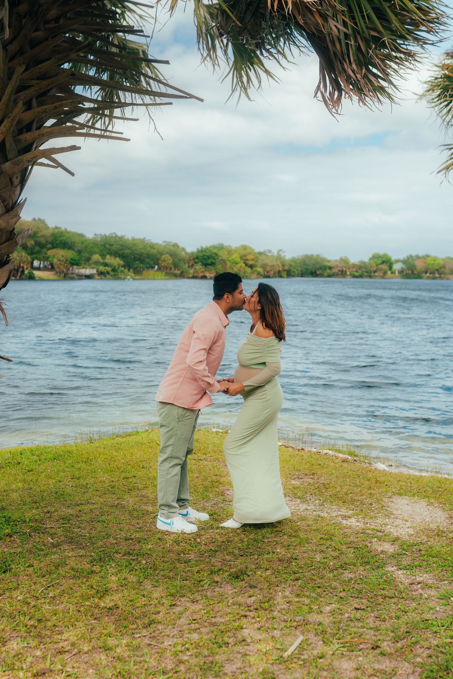 Couple sharing a kiss by a lakeside under a palm tree, woman in flowing sage green maternity dress.