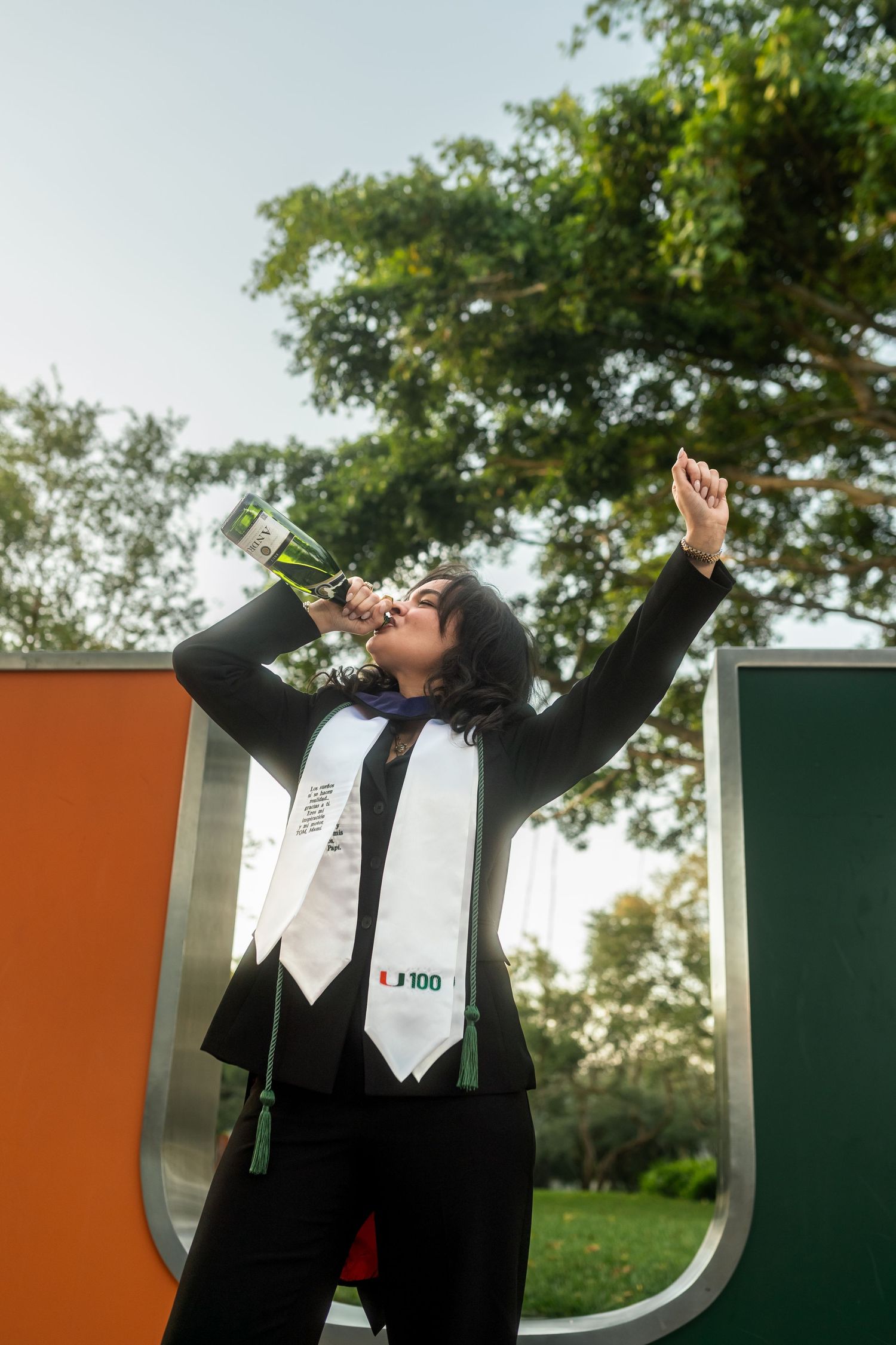 Graduate in black gown celebrates with champagne bottle raised near University of Miami U sign.