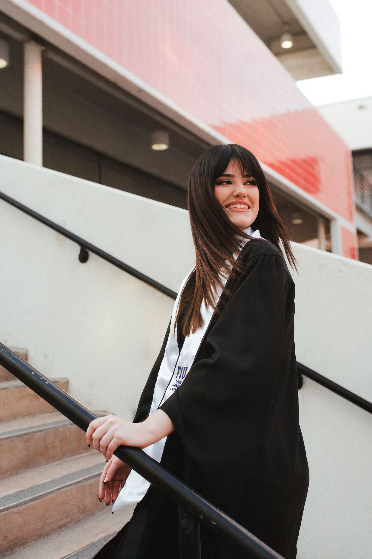 Smiling young woman in black graduation gown and sash poses on outdoor stairs at her university.