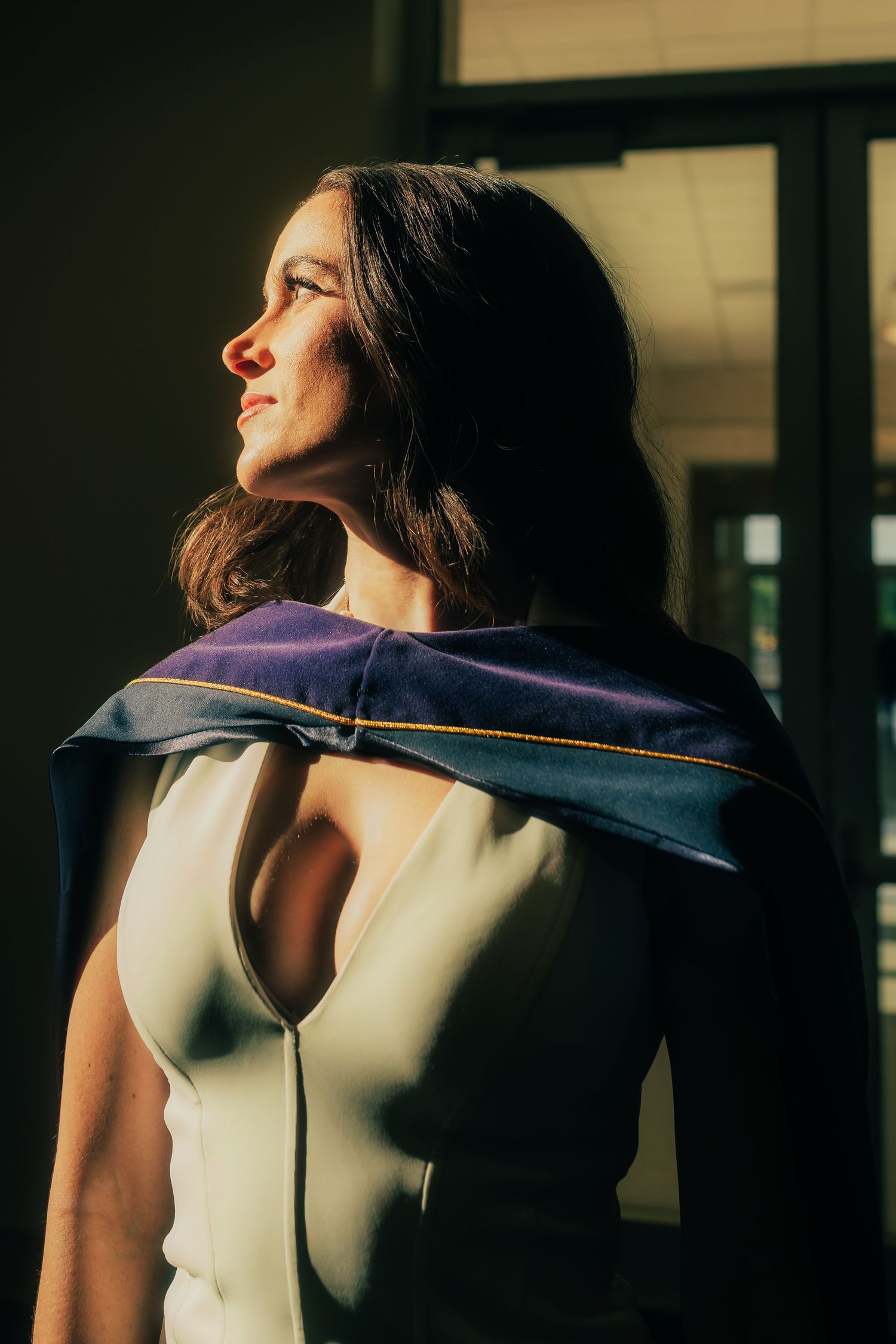 Woman in white dress with navy graduation hood poses in dramatic sunlight by window.