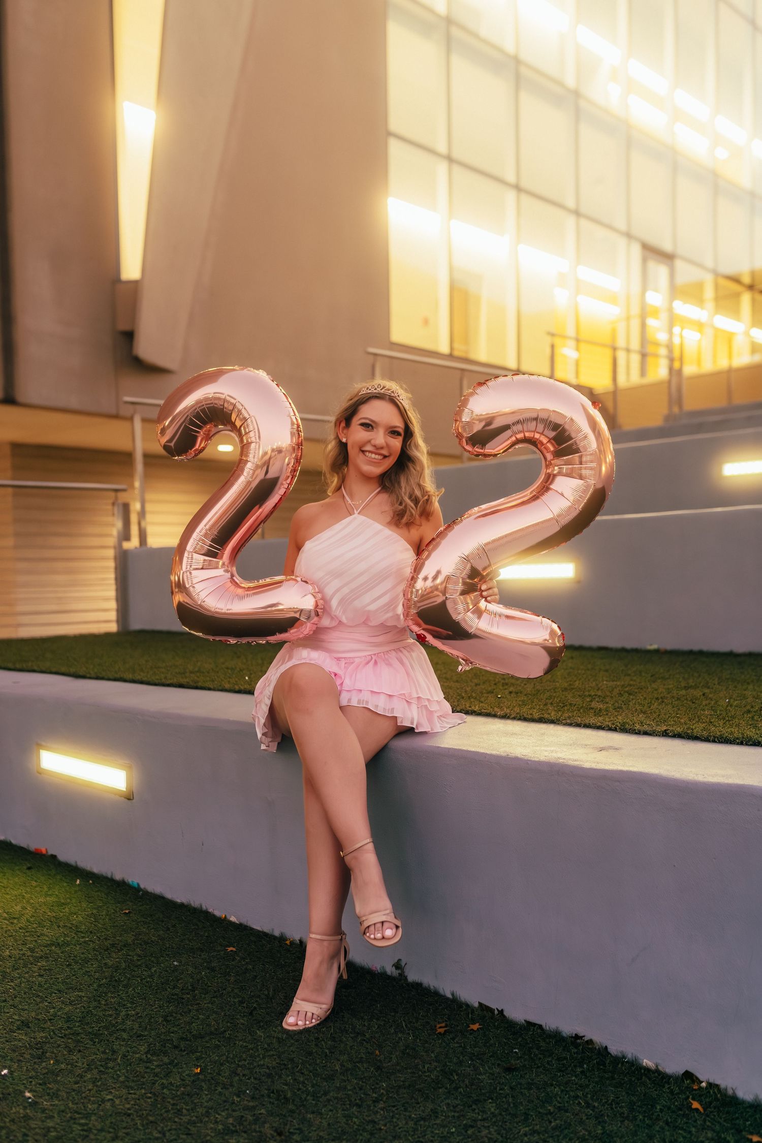 Smiling blonde woman in pink dress sitting outdoors holding rose gold number 2 balloons celebrating New Year 2022.