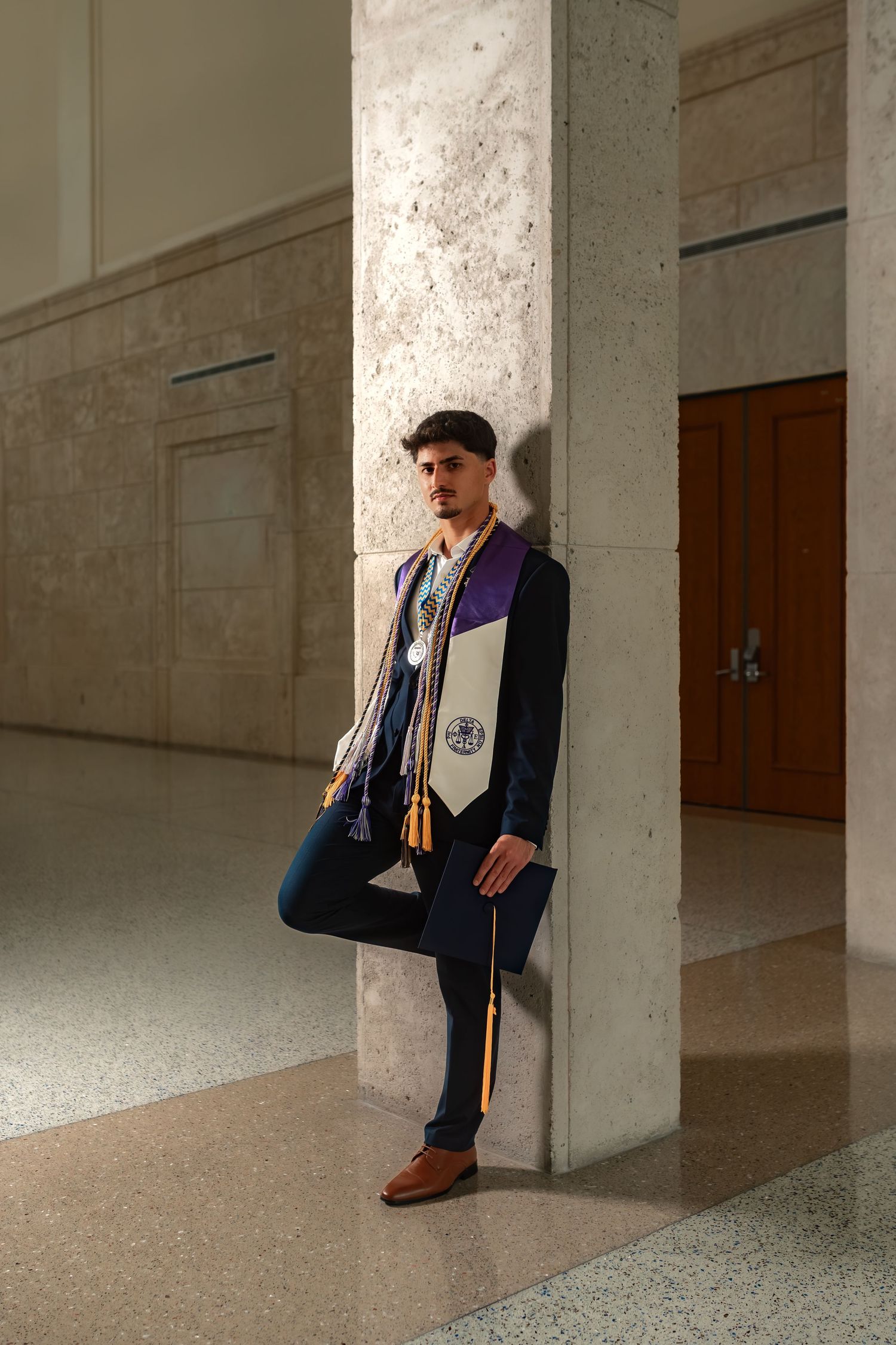 Male graduate in navy gown leans against marble column in elegant campus building.