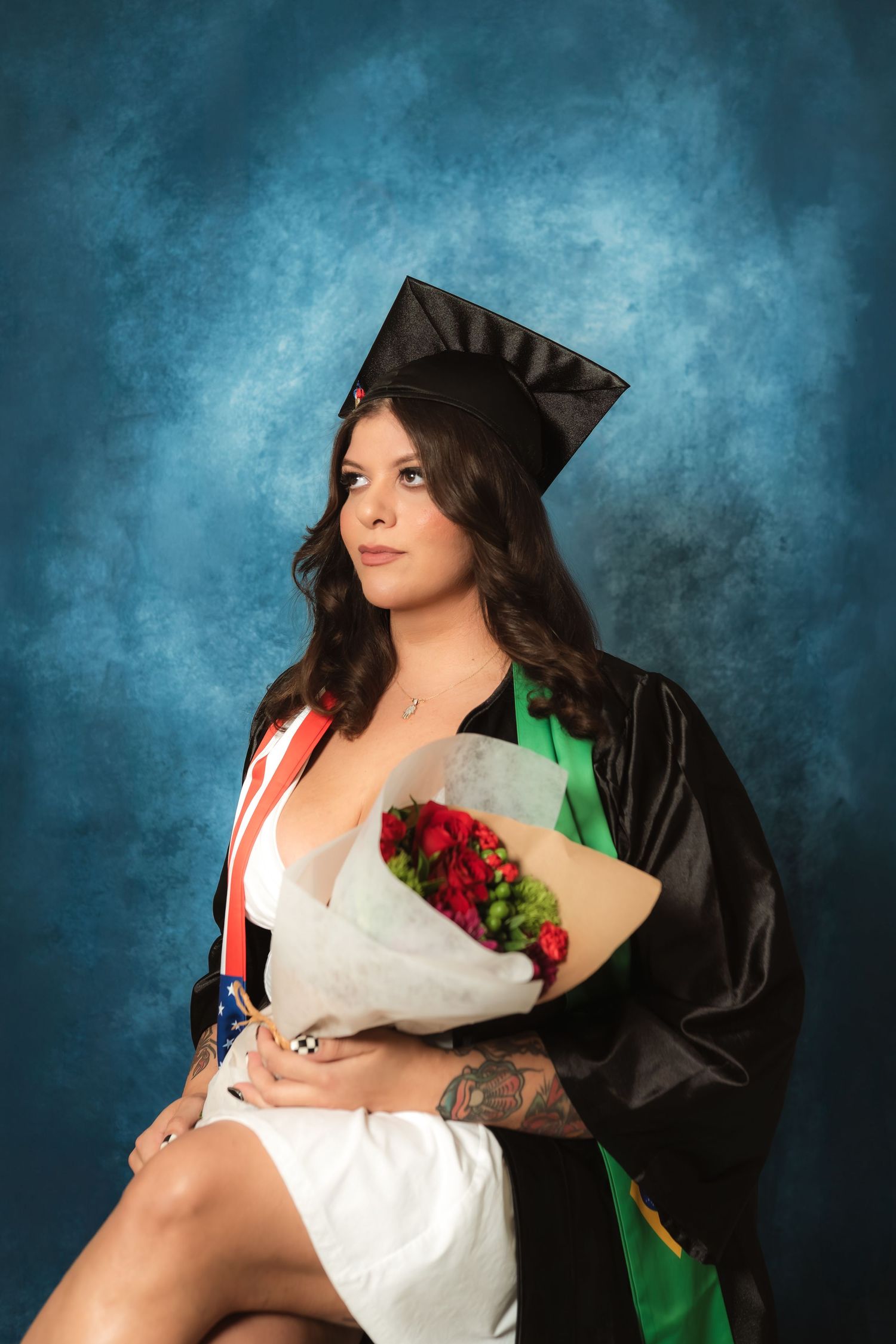 Young woman in graduation cap and gown holding red flowers, wearing a white dress with colorful sash, posed against blue backdrop.