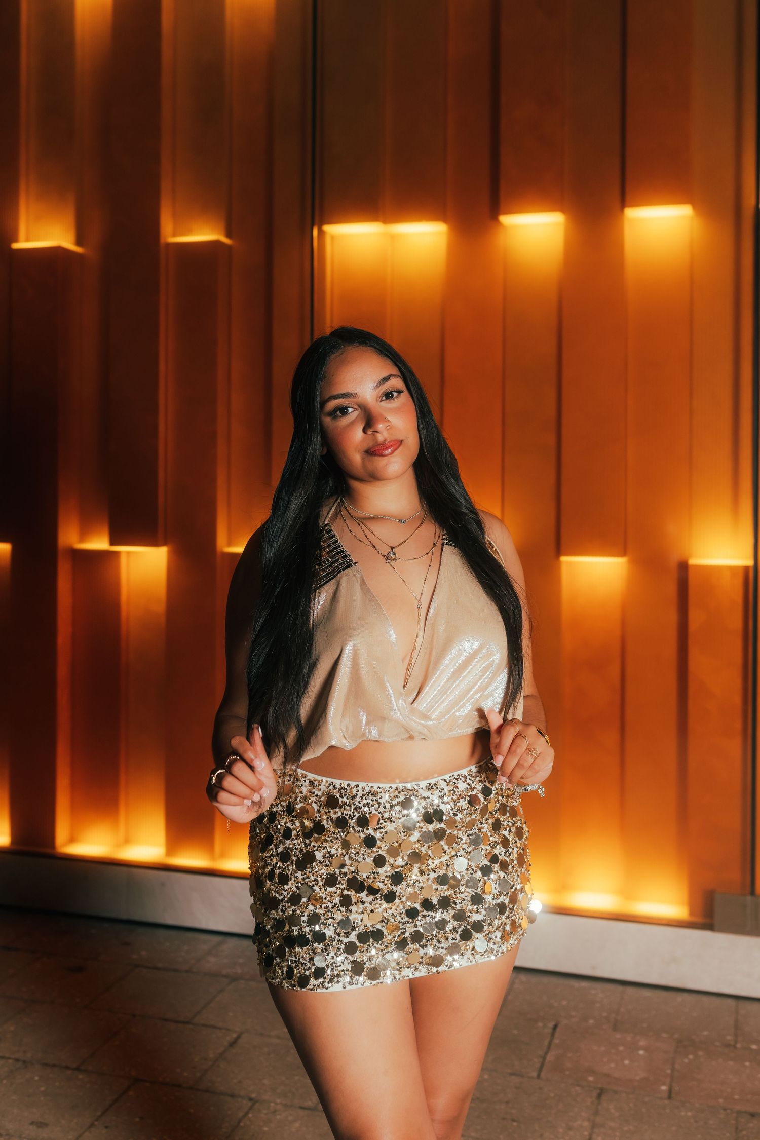 Young woman in sequin mini skirt and crop top poses against warm golden lit wall panels.
