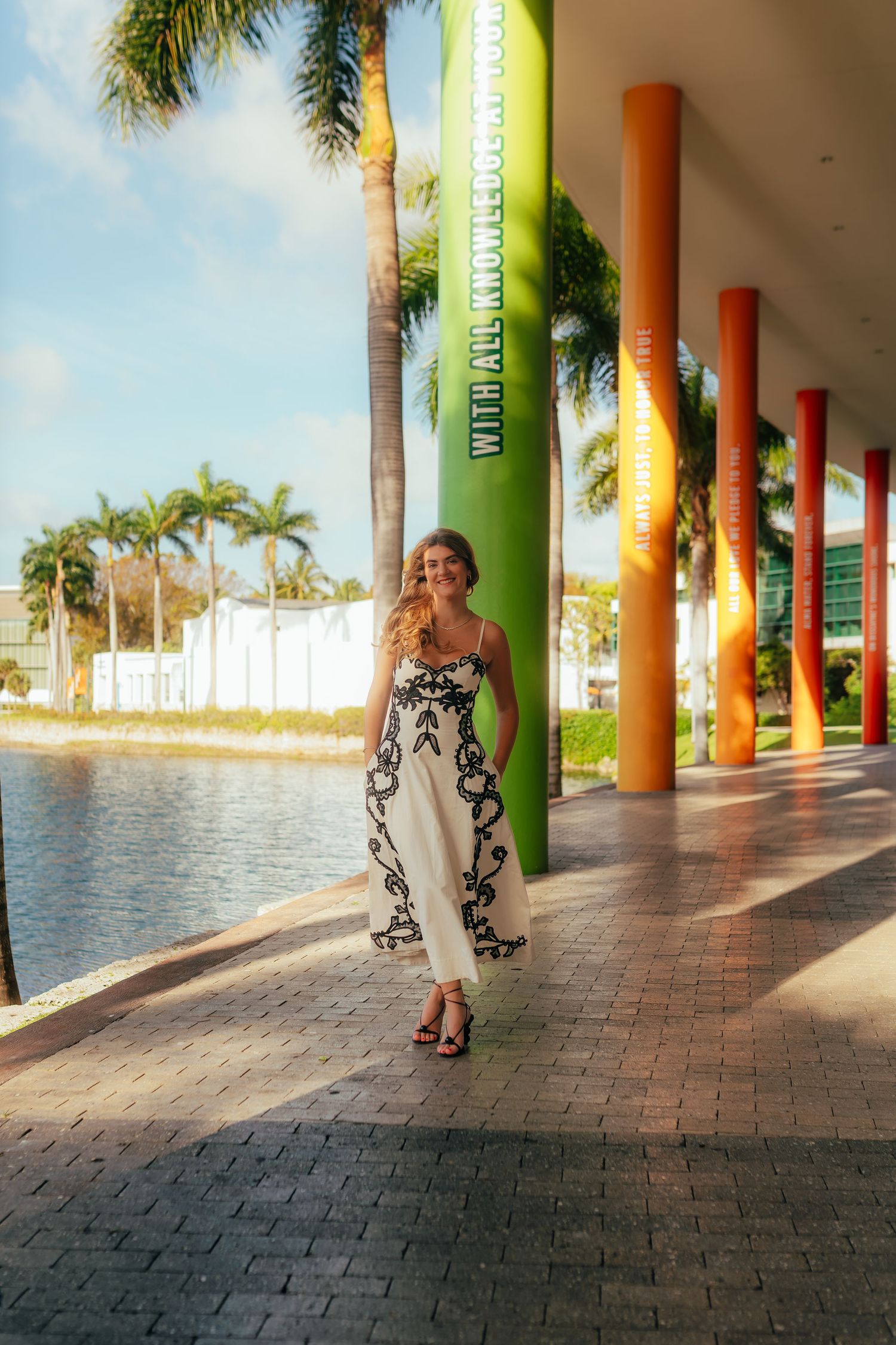 Woman in black and white maxi dress walks along colorful columned waterfront promenade with palm trees.