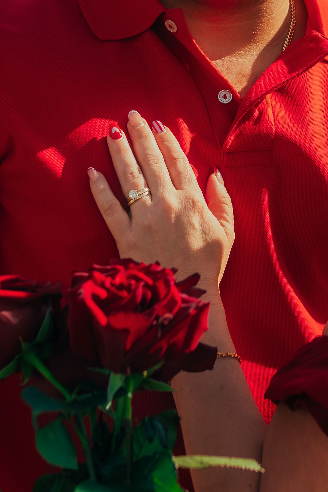 Close-up of woman's hand with diamond engagement ring resting on man's red shirt holding a rose.