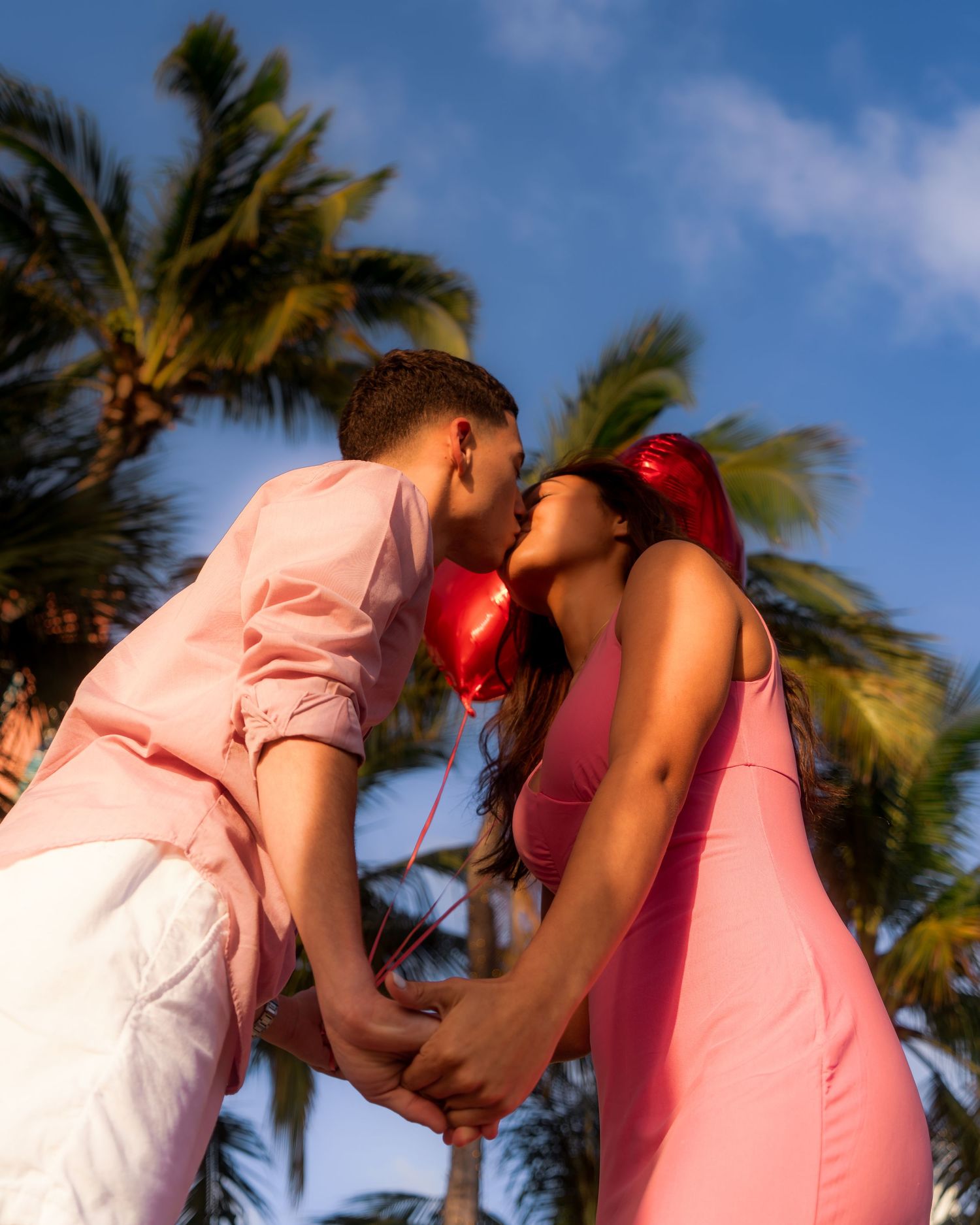 Young couple kissing under palm trees holding red heart balloons wearing matching pink outfits.