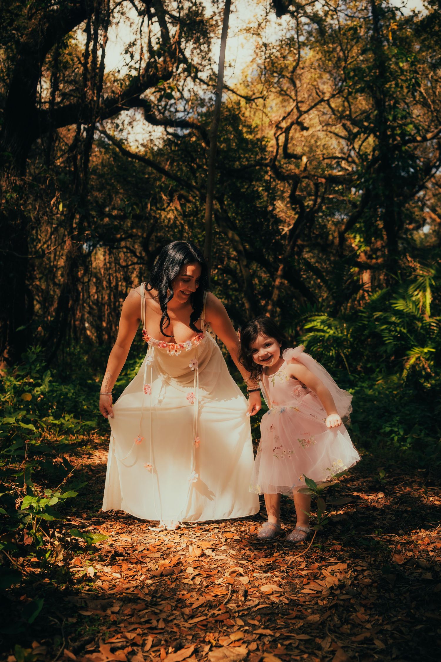 A woman in a white dress walks with a young girl in a pink tutu through a sunlit forest path covered in autumn leaves.