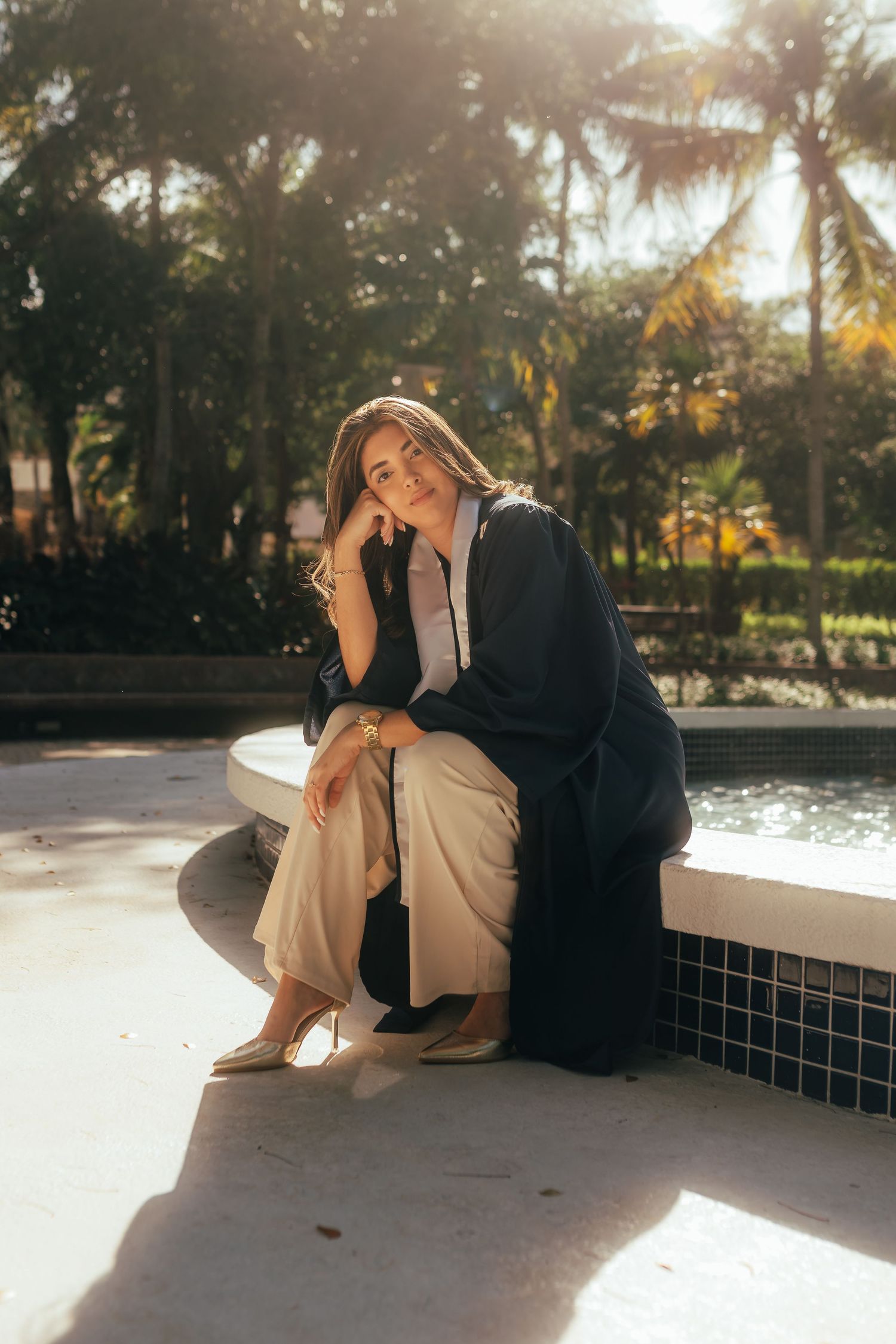 Woman in black graduation gown sits by fountain with palm trees in golden sunlight.