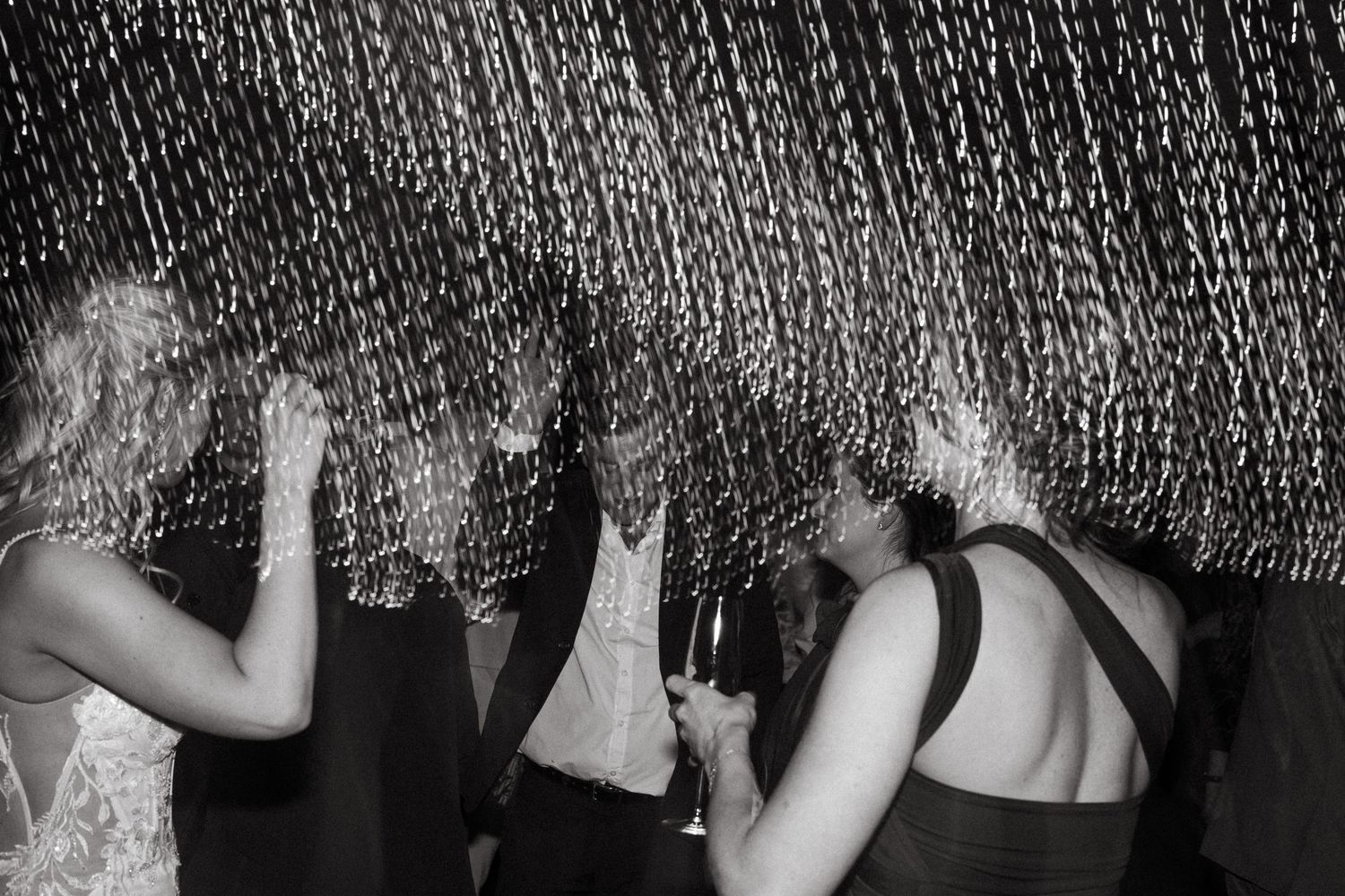 Black and white photo of people dancing at a party under shimmering tinsel curtain decorations.