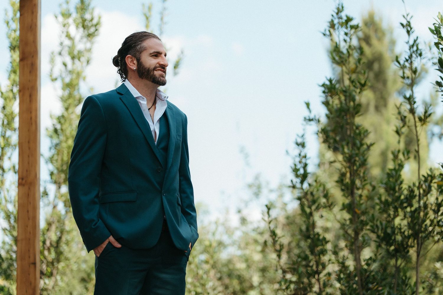 Man in teal suit with hair bun smiling outdoors among green trees on a bright day.