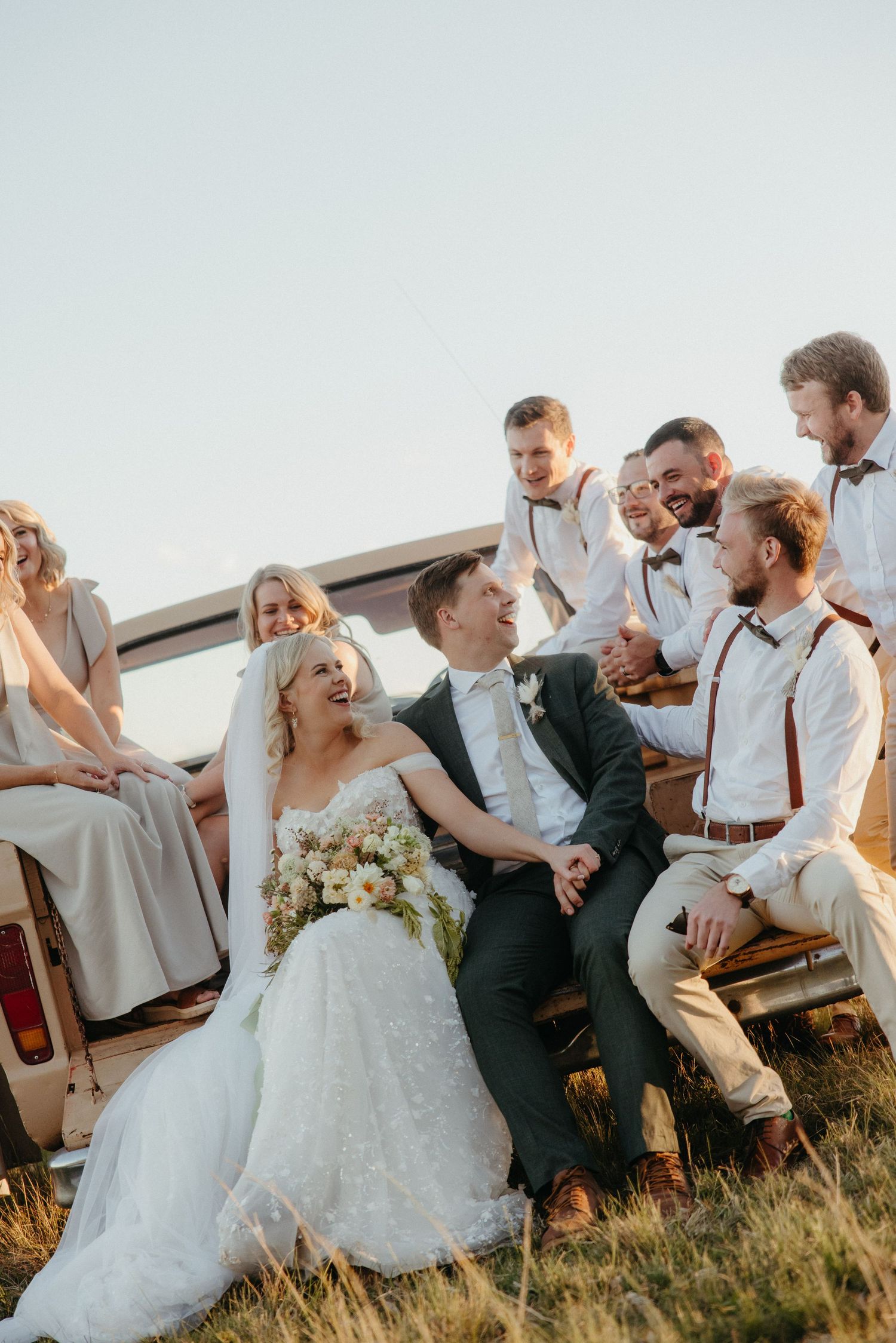 Bride and groom laugh together surrounded by wedding party seated on vintage vehicle in golden field.