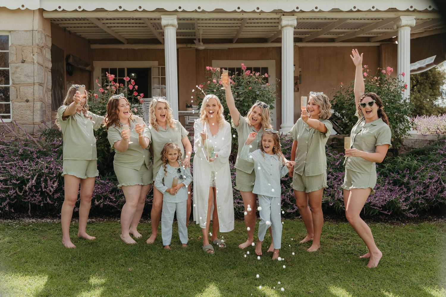 Bride in white lace robe celebrates with bridesmaids in sage green matching sets, tossing confetti outdoors.