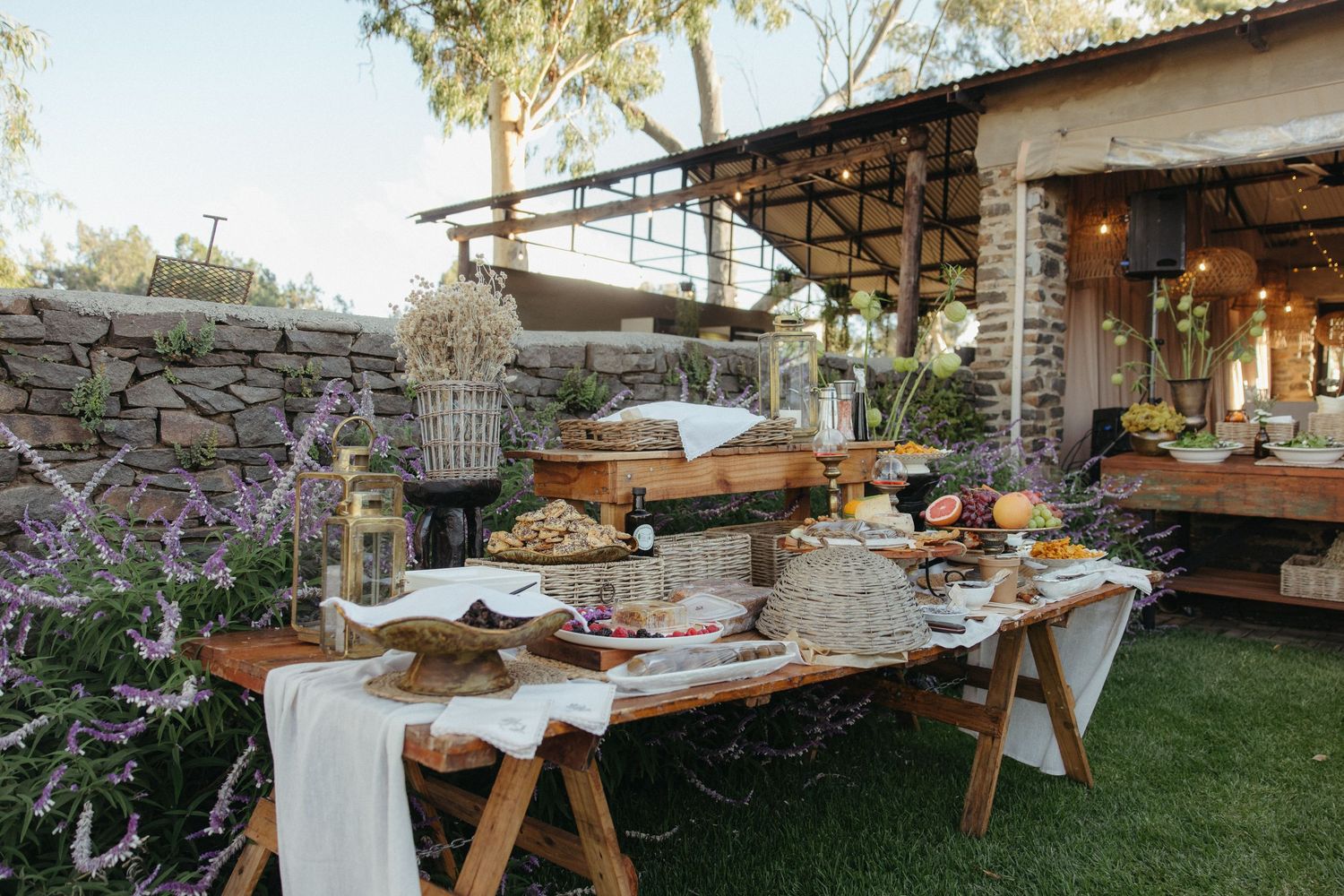 Rustic outdoor buffet table with food spreads, wooden furniture, string lights, and lavender plants at a garden event.