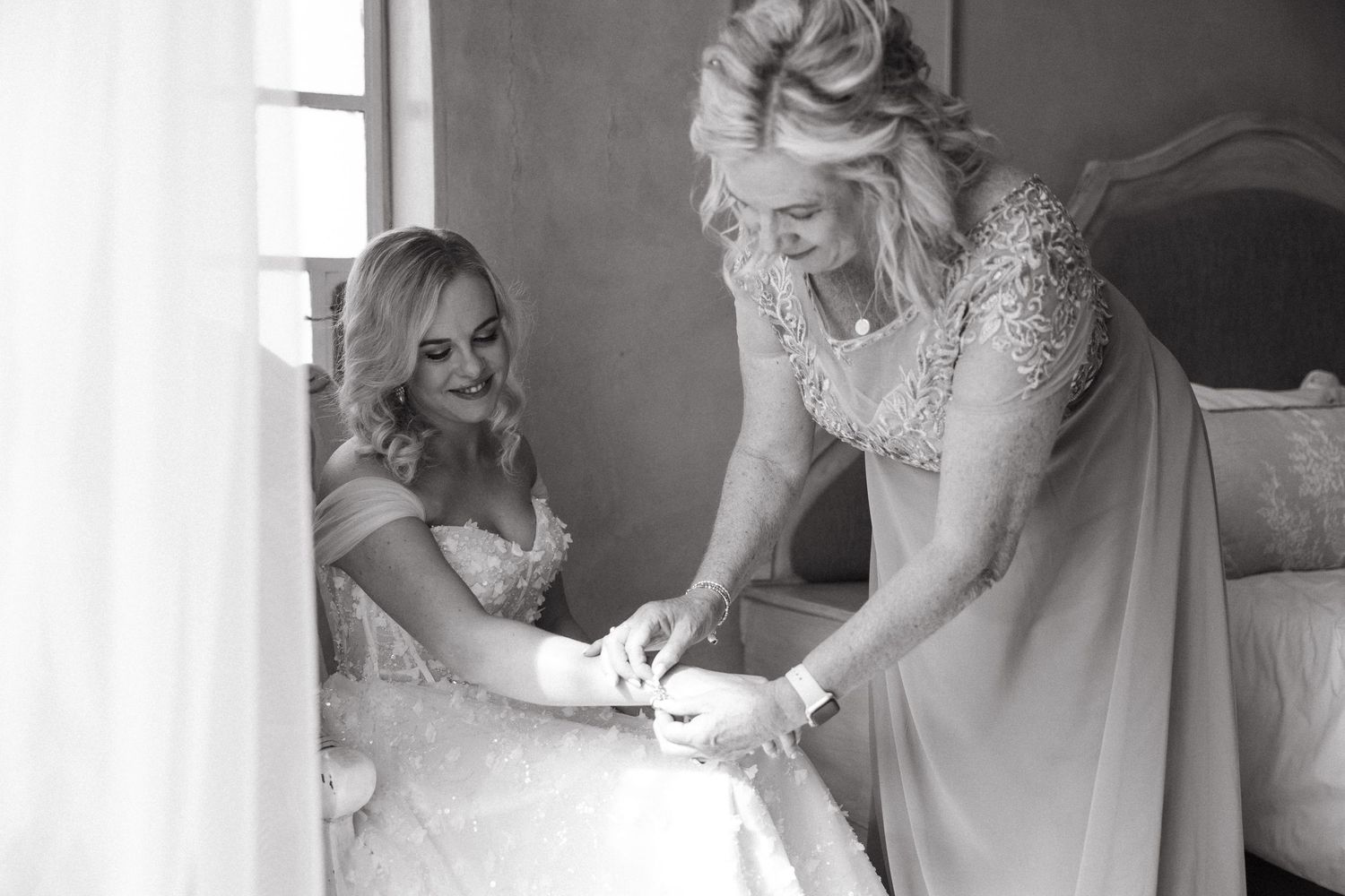 Black and white photo of bride in wedding gown being helped by woman in lace dress near window.