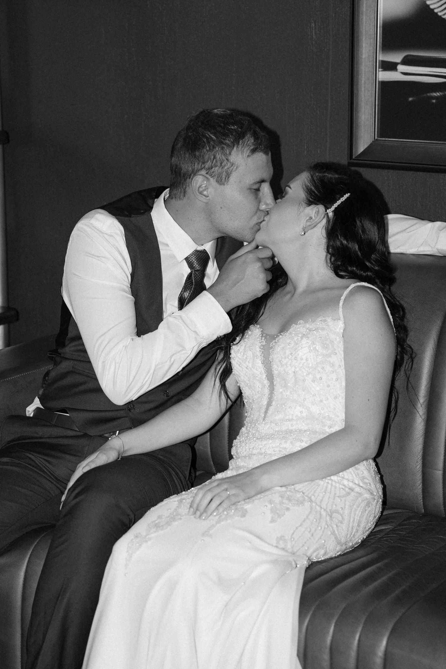 Black and white photo of a bride and groom sharing a romantic kiss while seated on a leather sofa at their wedding.