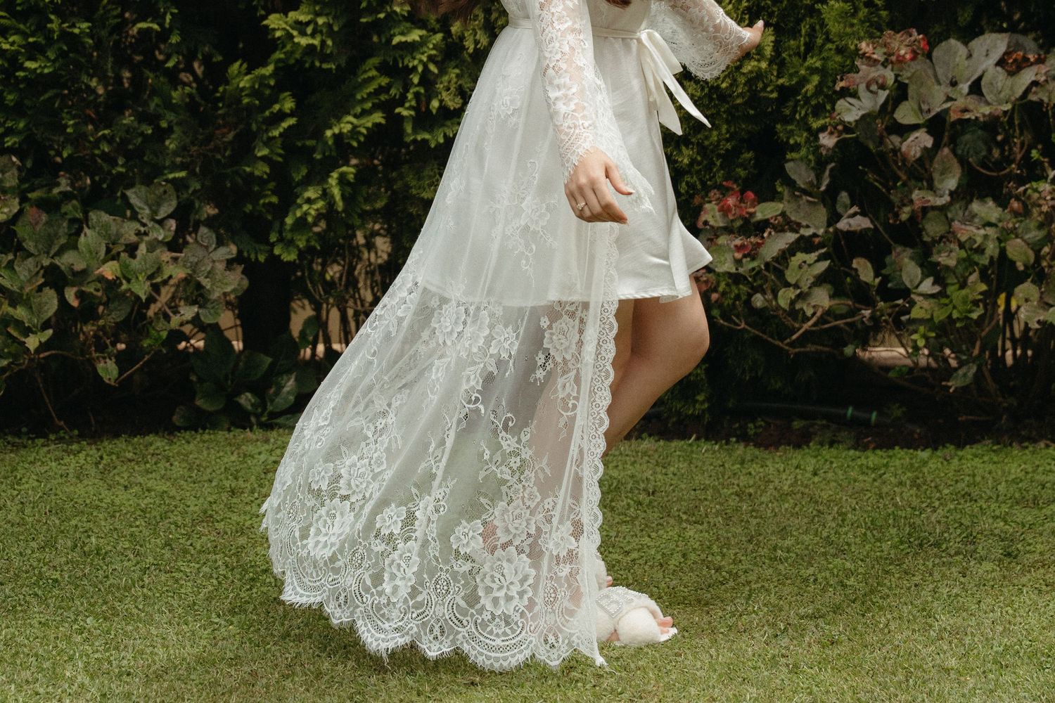 Bride in a flowing white lace wedding gown walking on green grass with lush garden backdrop.