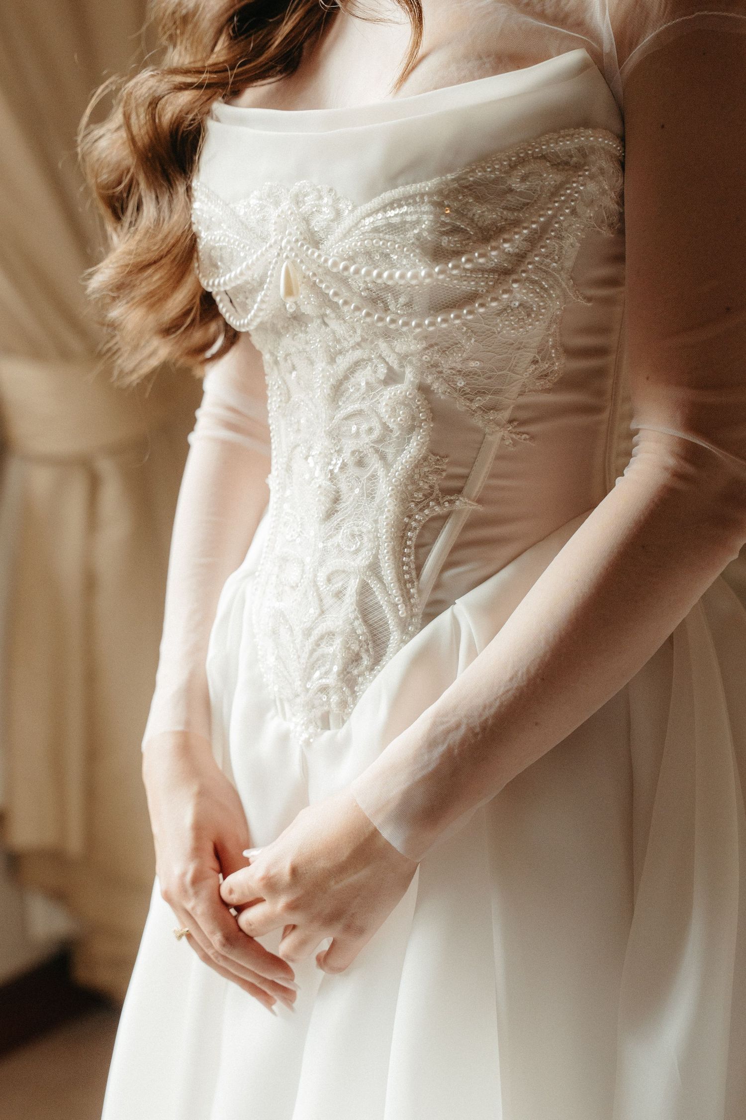 Bride in elegant white lace corset wedding gown with sheer sleeves, hands clasped, soft natural light.