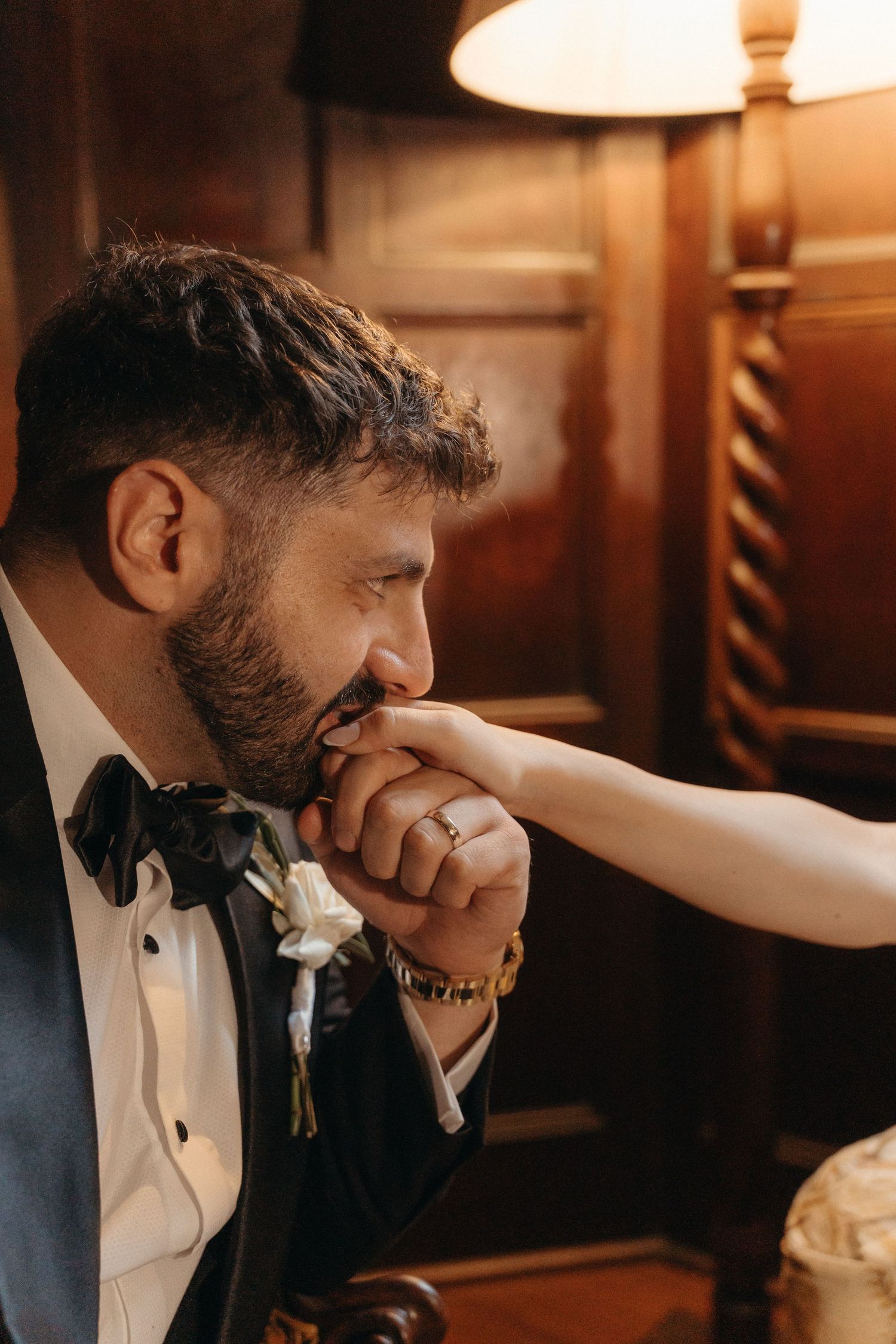 Groom in tuxedo kissing bride's hand tenderly, wearing a gold watch and boutonniere in warm romantic lighting for Glenshiel wedding photos