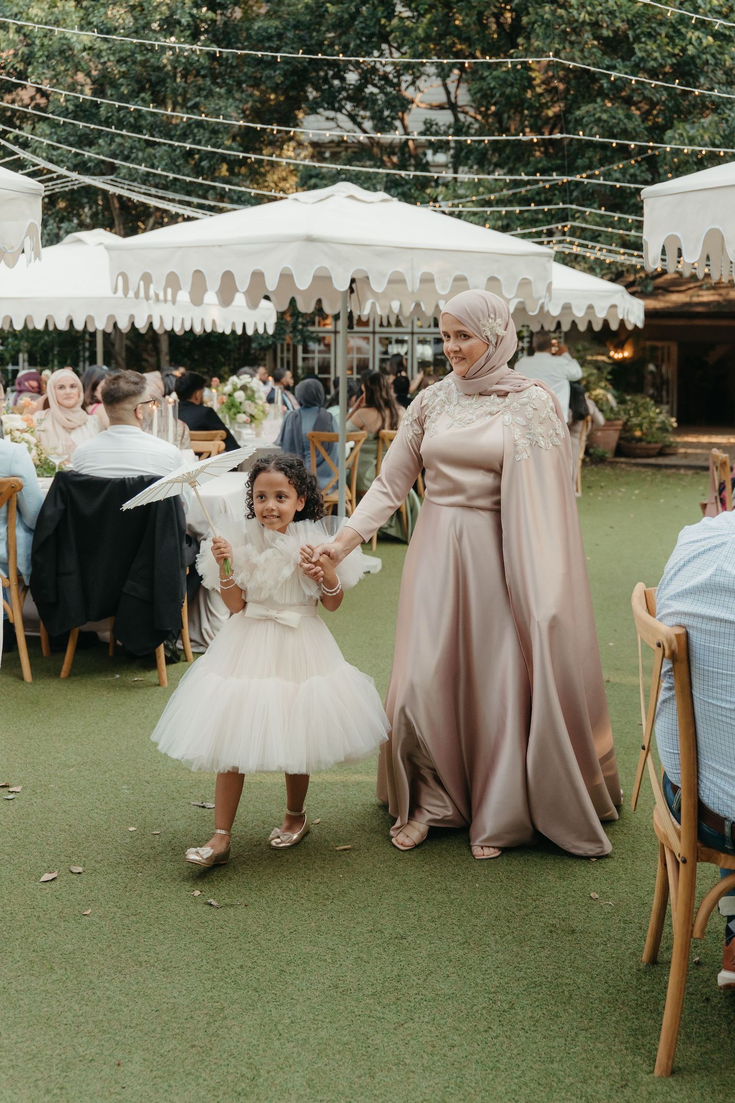 A woman in a hijab and elegant gown walks hand-in-hand with a little girl in a white tutu at an outdoor wedding.