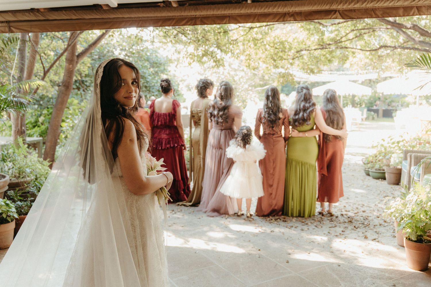 Bride in white gown and veil holds bouquet while bridesmaids in colorful mismatched dresses stand together outdoors.