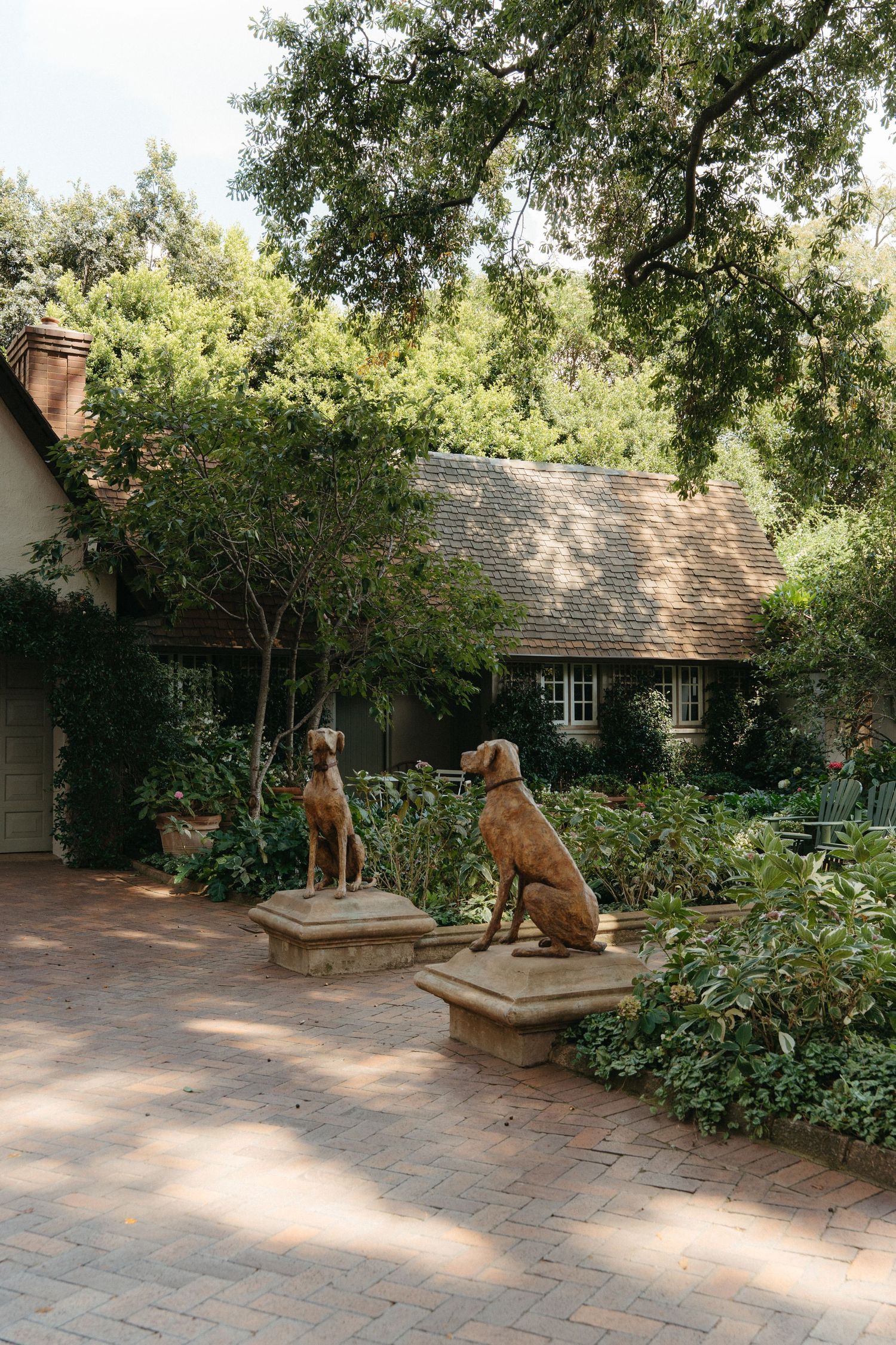 Two bronze dog statues on stone pedestals adorn a brick patio outside a charming cottage surrounded by lush greenery in Johannesburg