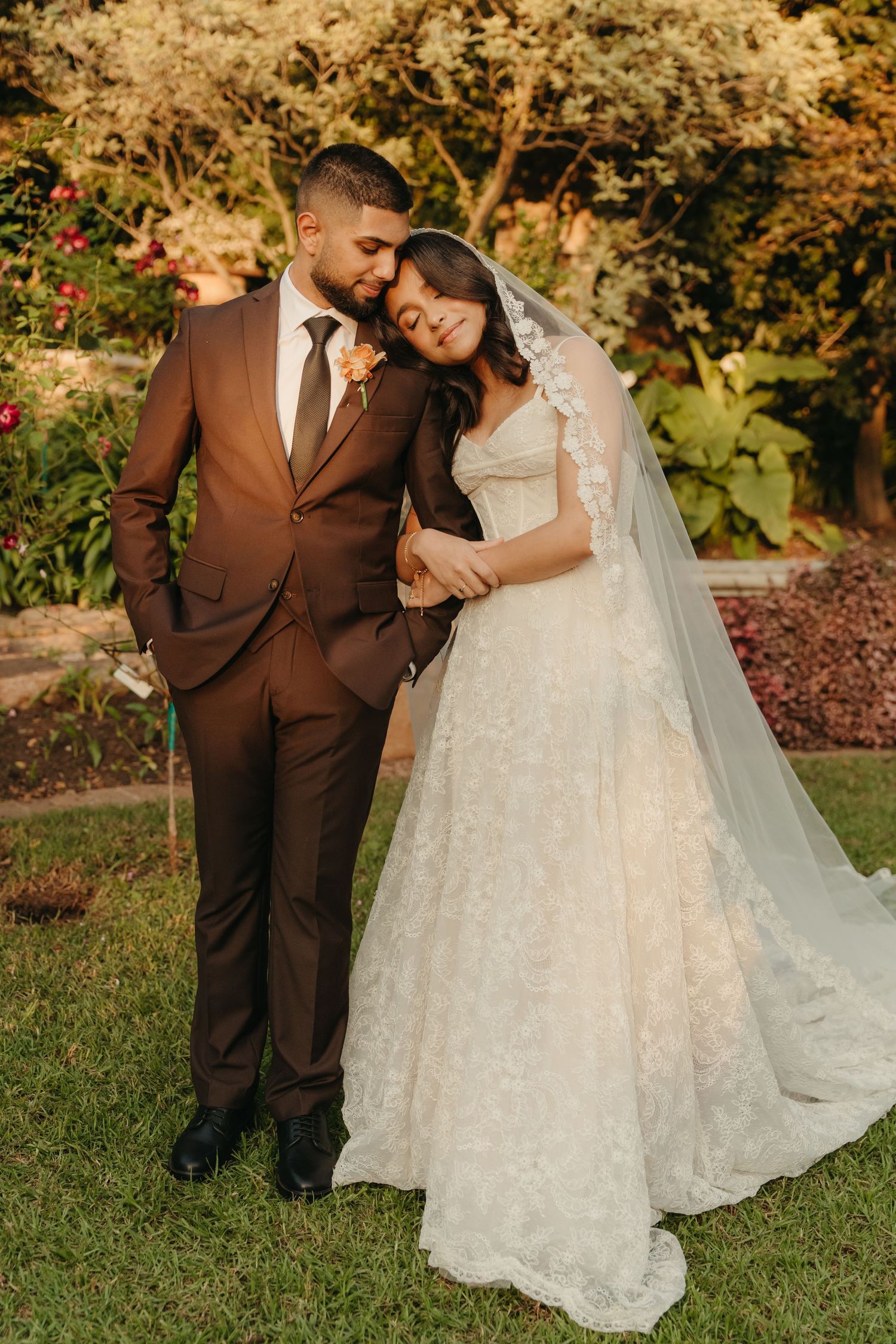 Bride in lace gown and veil leans on groom in brown suit, posing together in a lush garden setting.