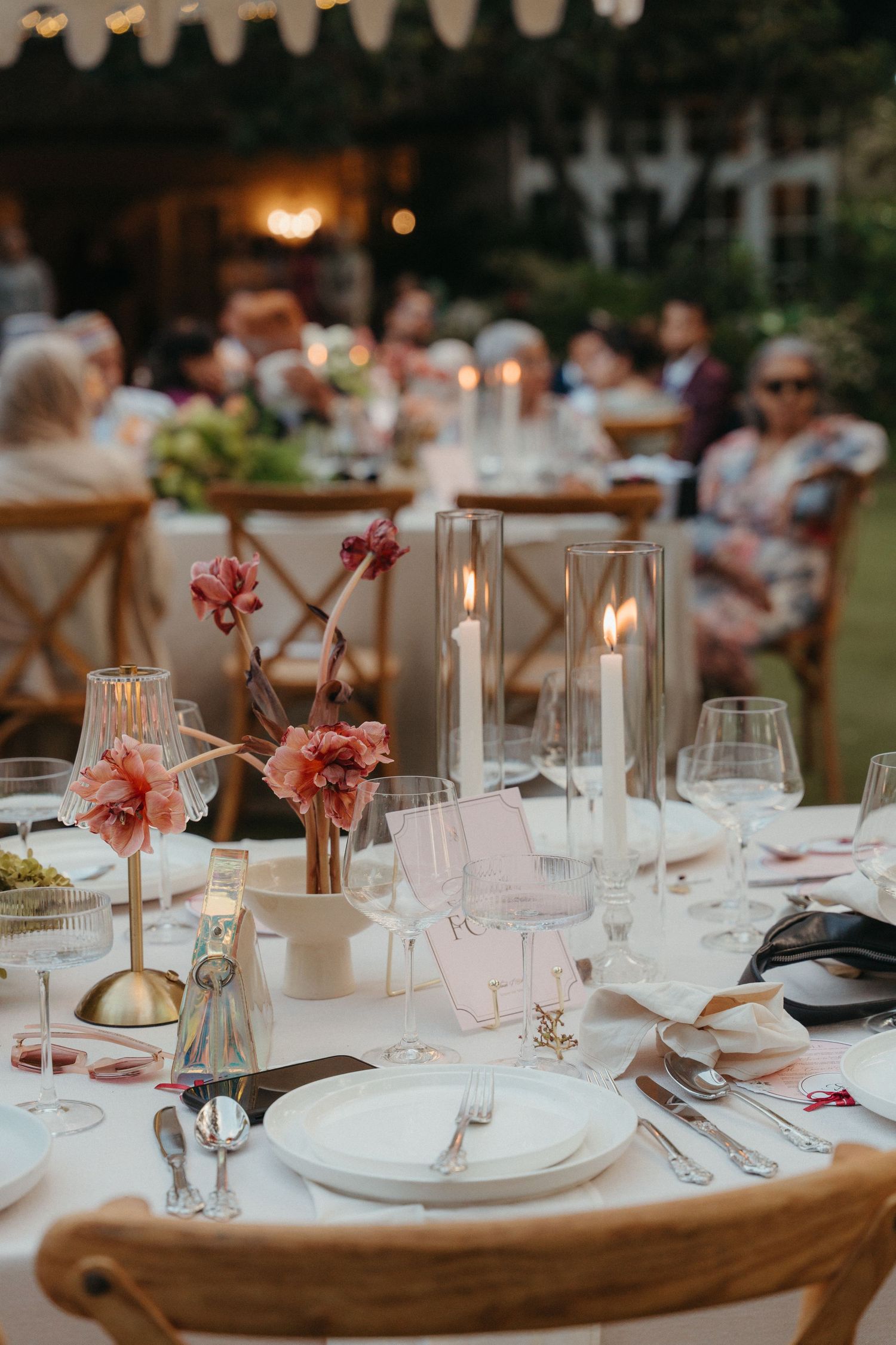 Elegant outdoor wedding reception table with candles, pink floral centerpieces, white plates, and glassware at dusk.