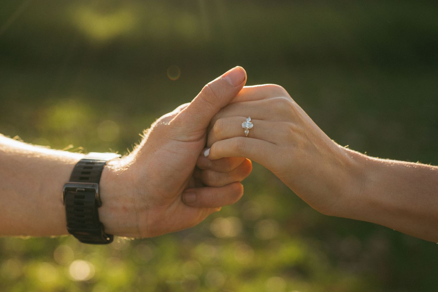 Two hands tenderly touch while one person wears a black watch and the other wears an engagement ring in soft sunlight.