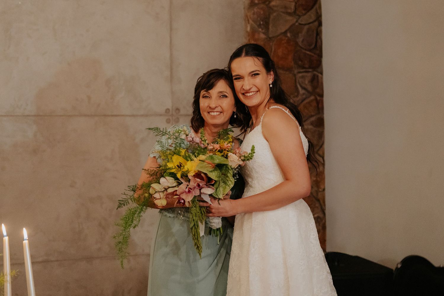 Two people in wedding attire embrace while standing in front of a stone wall with natural light.