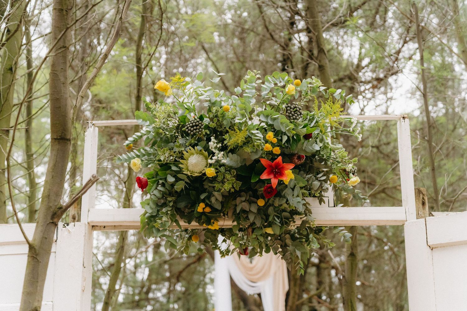 Vibrant floral arrangement with colorful wildflowers on a rustic white bench in a wooded setting.