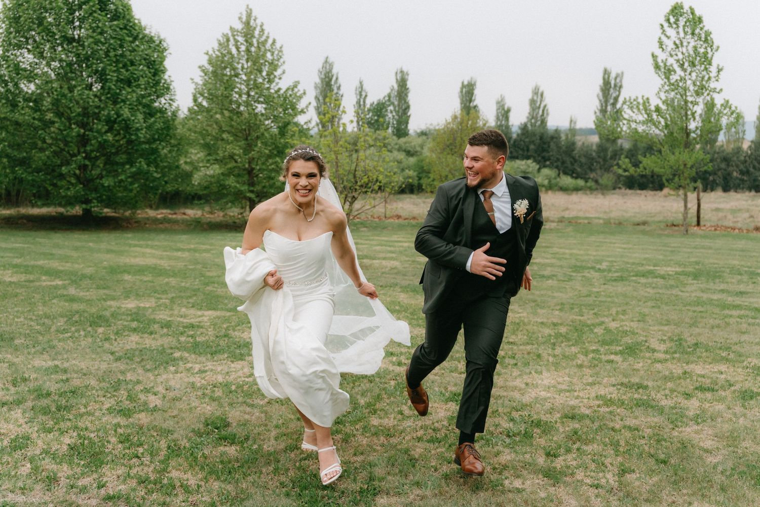 Newlyweds run playfully through a green field, with the bride's dress flowing in the wind.