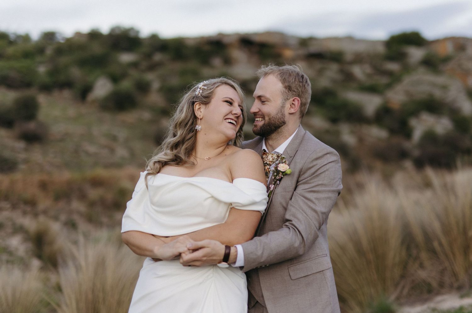 A couple shares a romantic embrace in an outdoor setting with desert hills and dry grass in the background.