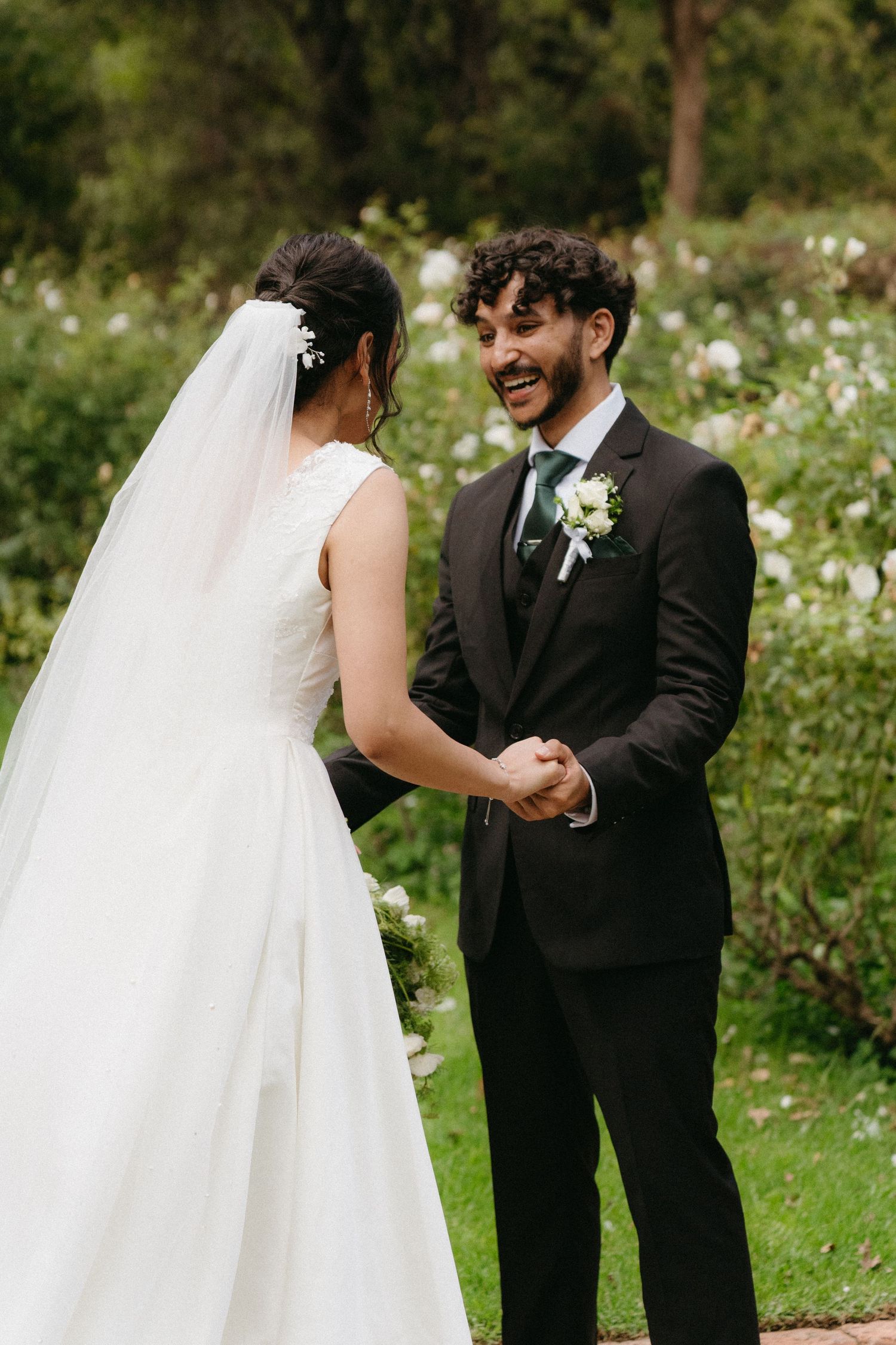 A bride and groom share a joyful first look moment in a lush garden setting with white flowers in the background.