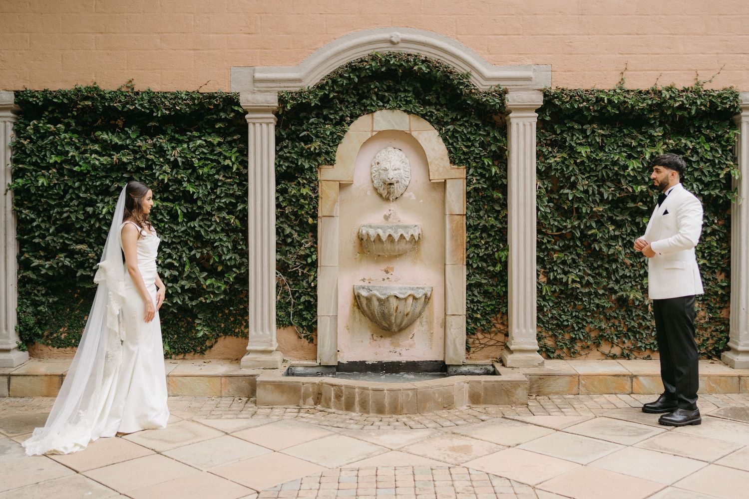 A couple standing by an elegant stone fountain with ivy-covered wall during an outdoor wedding ceremony.