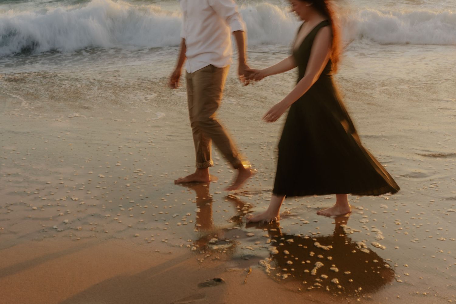 Couple walking along beach shoreline at sunset with waves gently rolling in.