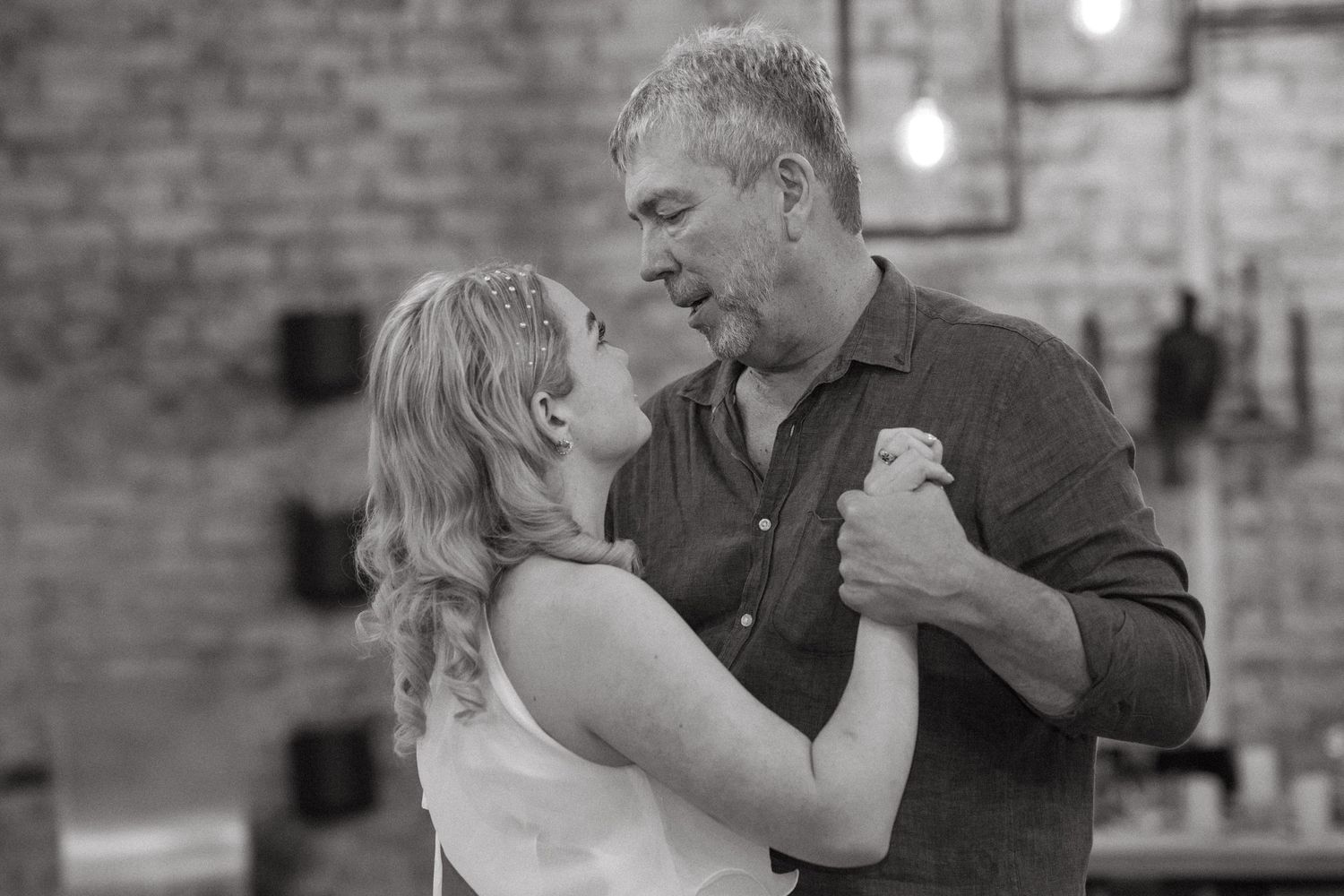 Two people share an intimate dance together in black and white against a brick wall backdrop.