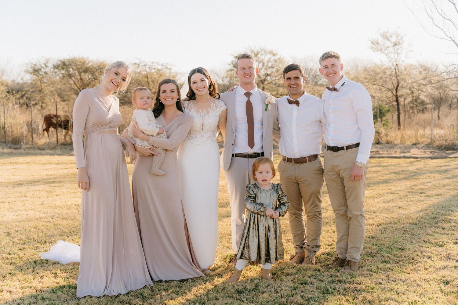 Wedding group photo with attendants wearing neutral colors standing together in a grassy outdoor setting.